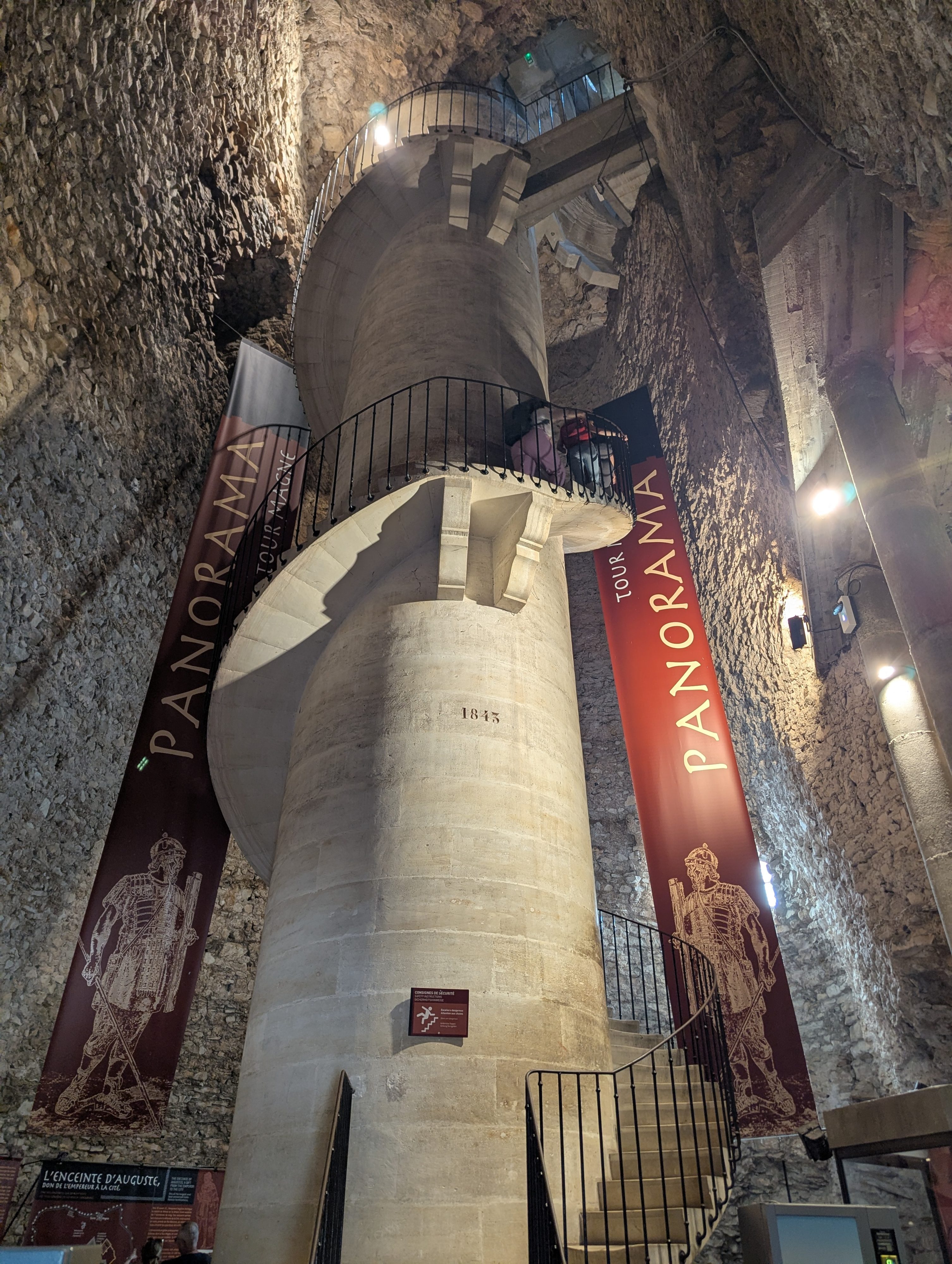 Looking up inside the Tour Magne at a curving staircase winding around a stone column, with red Panorama banners hanging on the rough stone walls