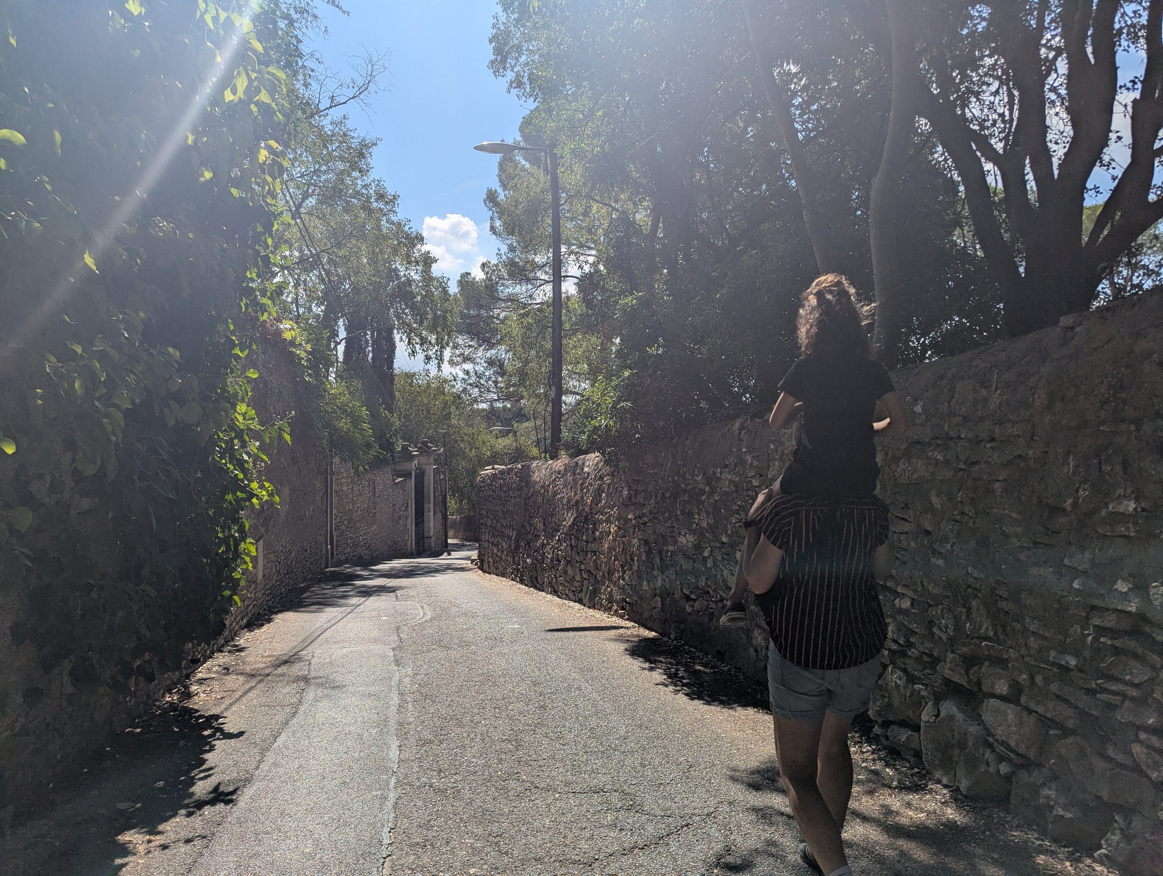 A parent walking up a sunlit lane between old stone walls and trees with Remi riding on their shoulders