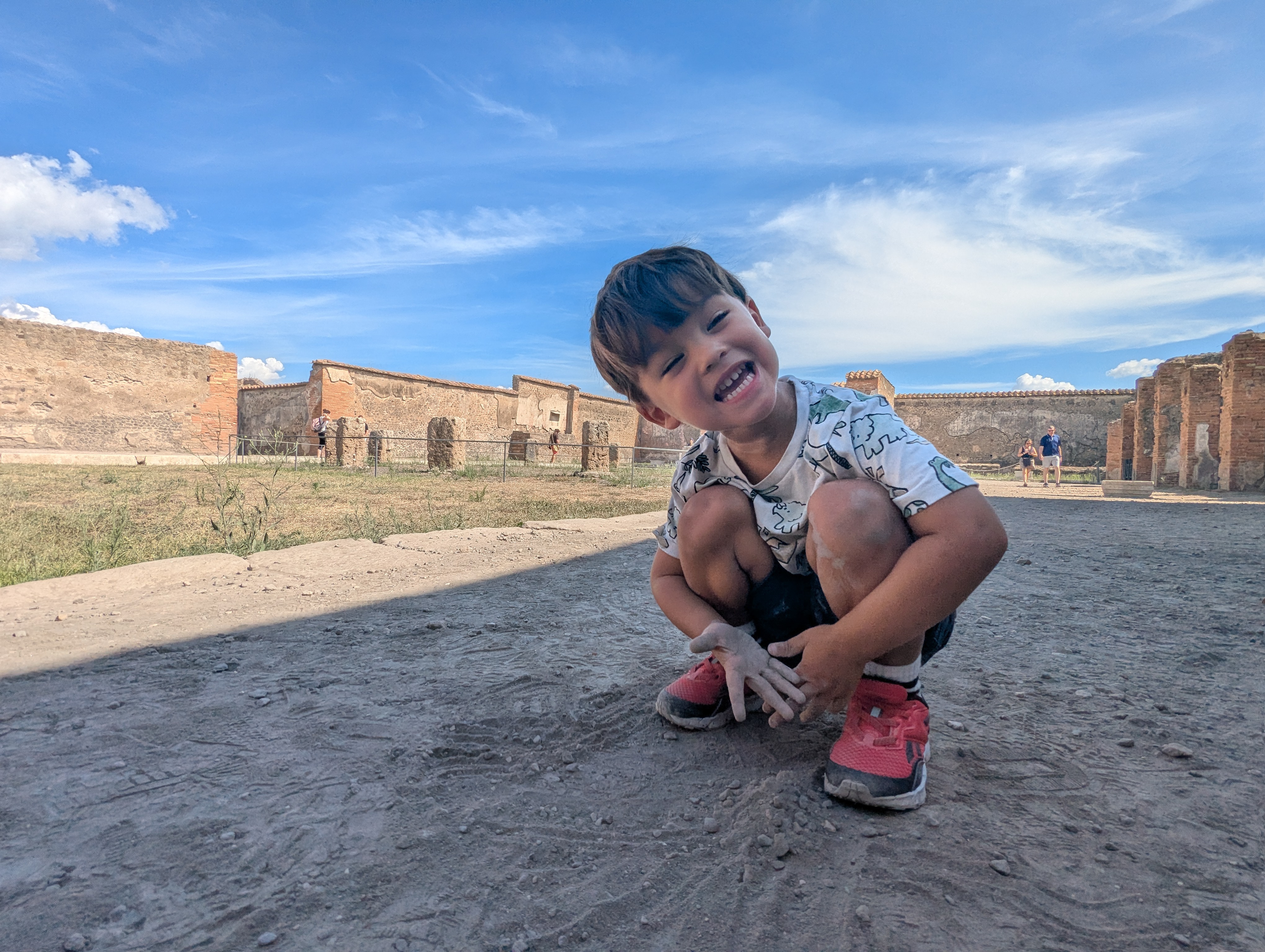 Arlo crouched in the dirt digging with both hands, grinning, ruins in the background