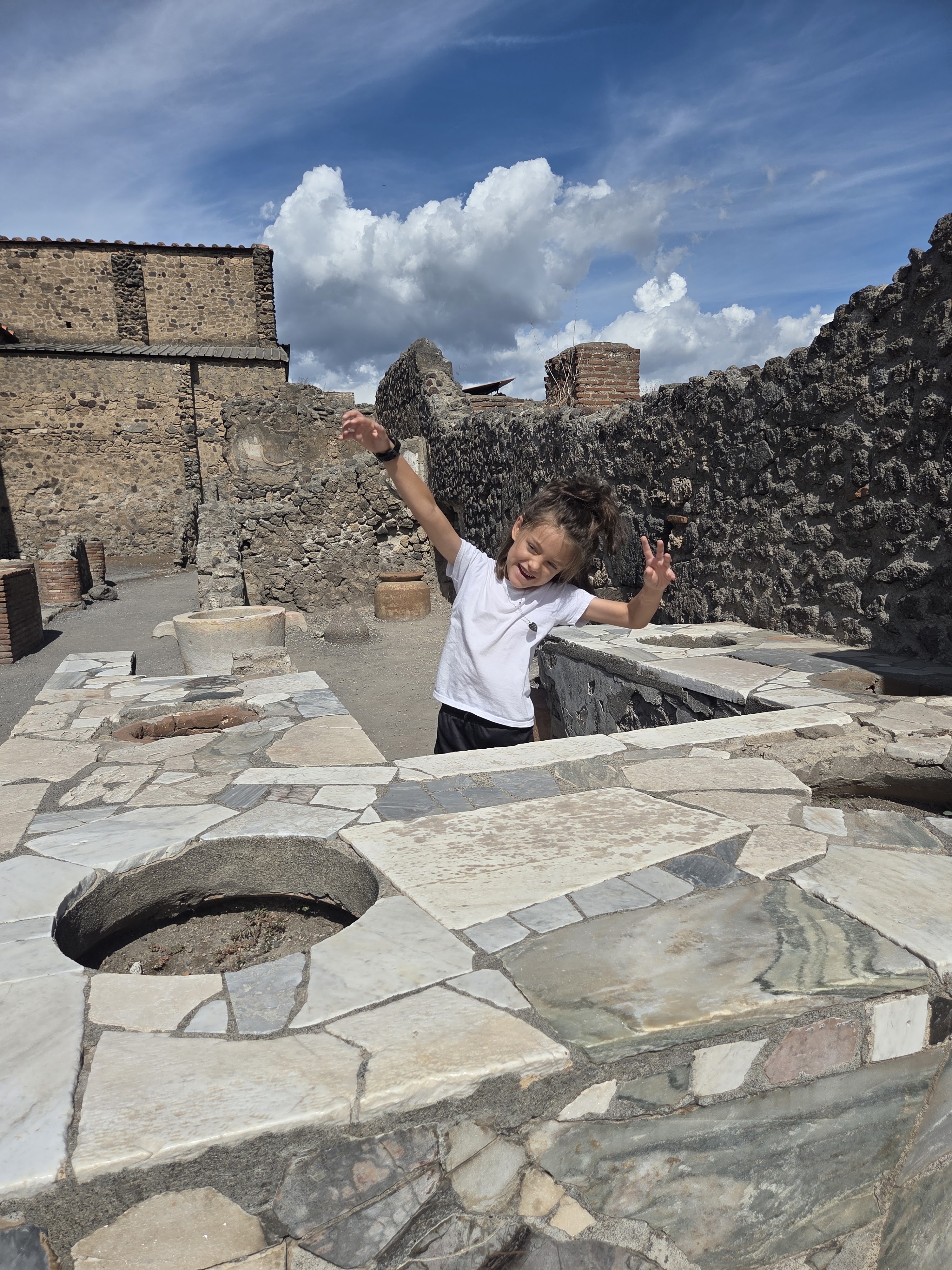 Arlo cheering with both arms raised at a stone thermopolium counter set with round jar holes