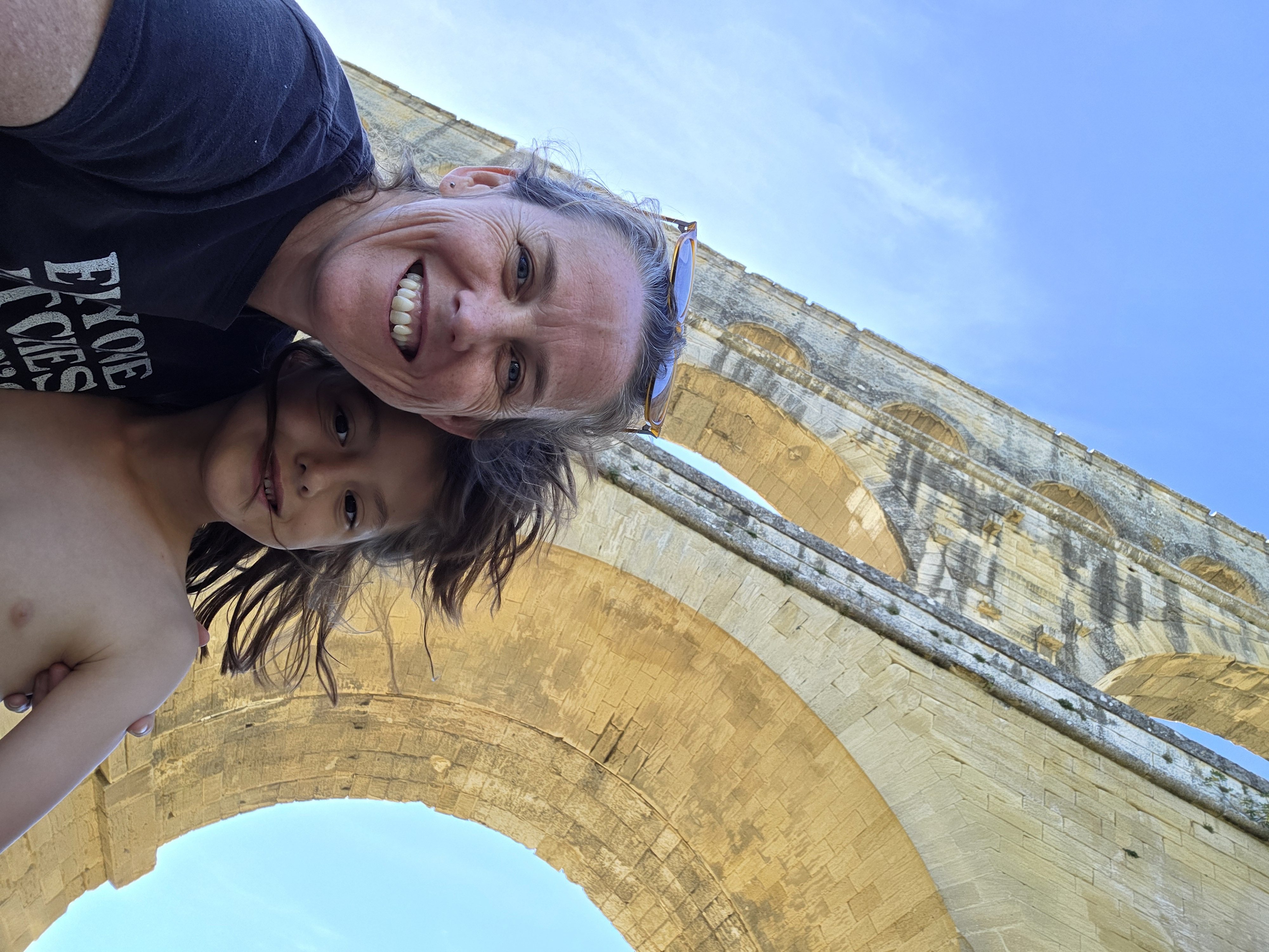 Dani and Remi taking a selfie from below with the massive stone arches of the Pont du Gard filling the sky behind them