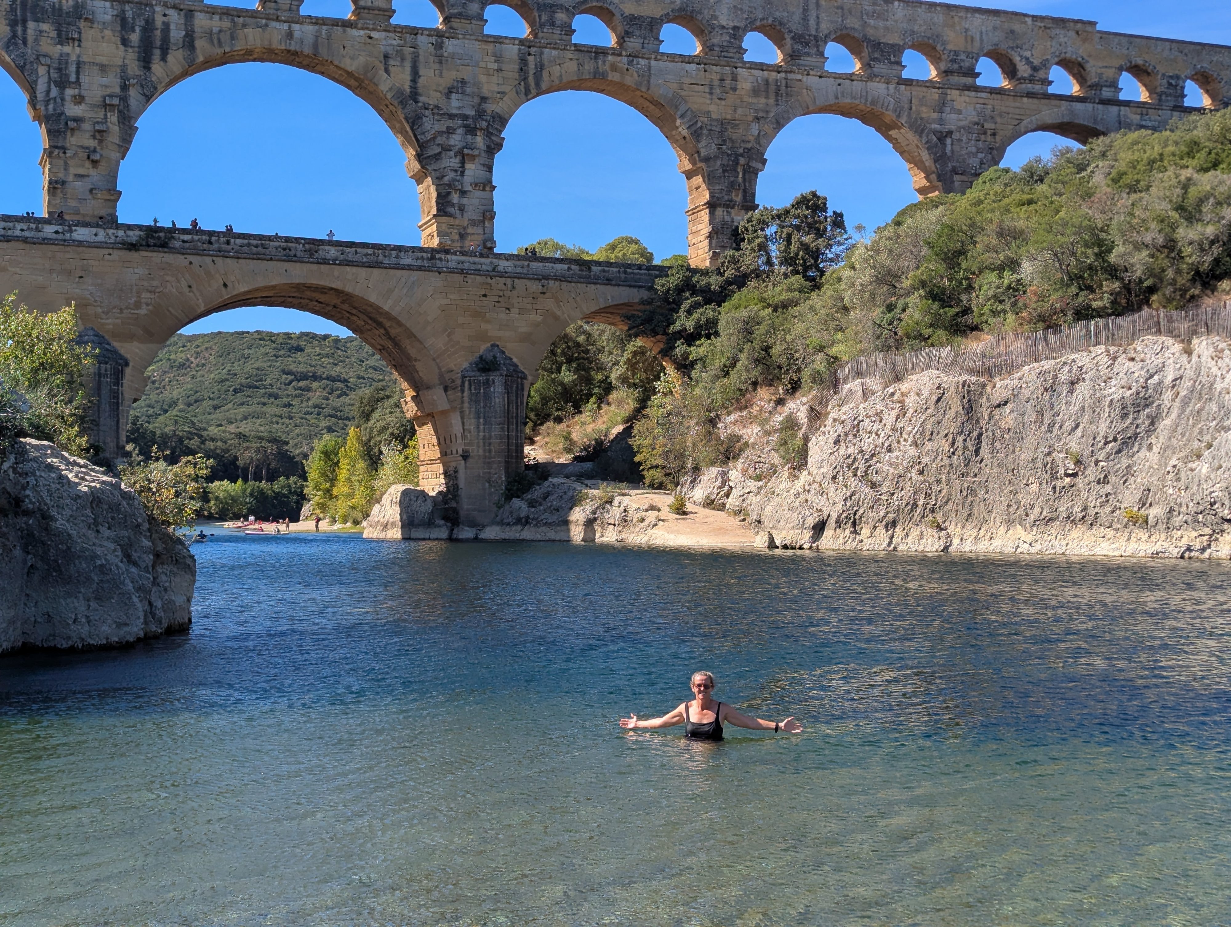 Dani standing waist-deep in the clear blue-green Gardon river with arms spread wide, the full span of the Pont du Gard aqueduct rising behind her