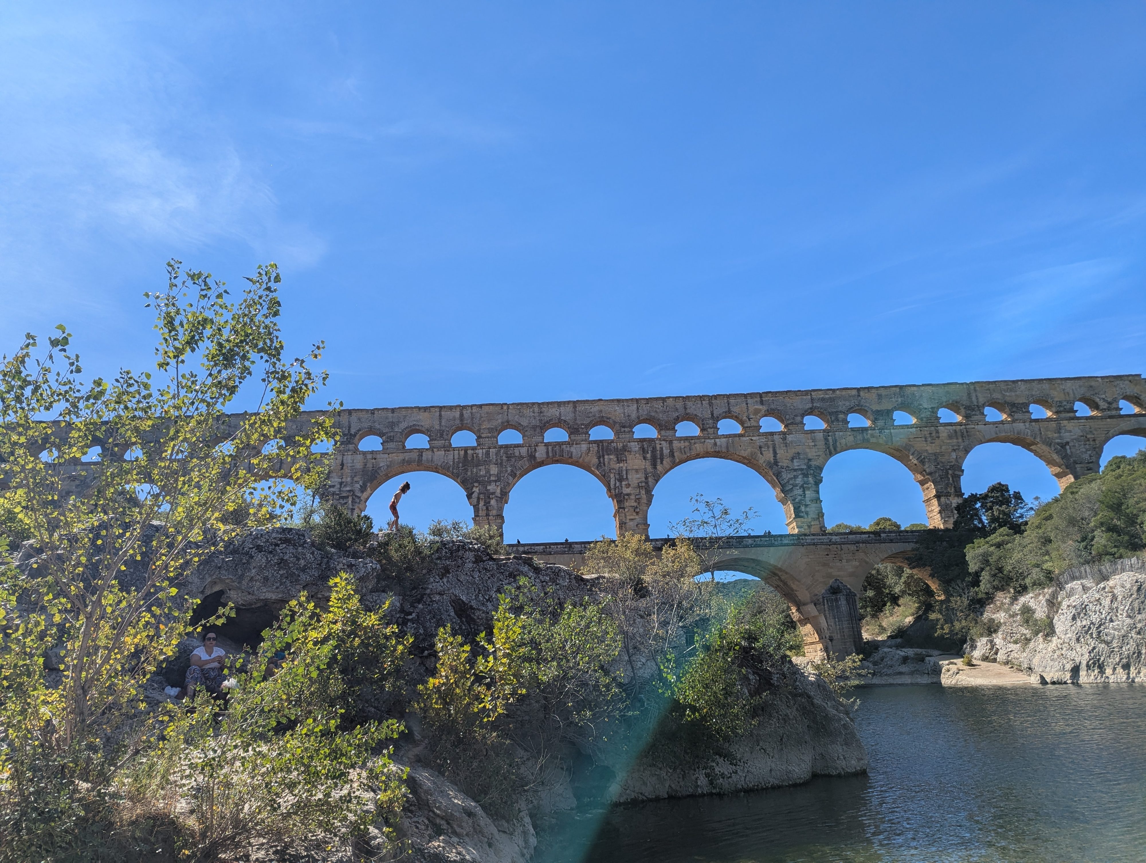 The Pont du Gard from a distance, all three tiers visible, with rocky outcrops and green trees framing the aqueduct and the river below