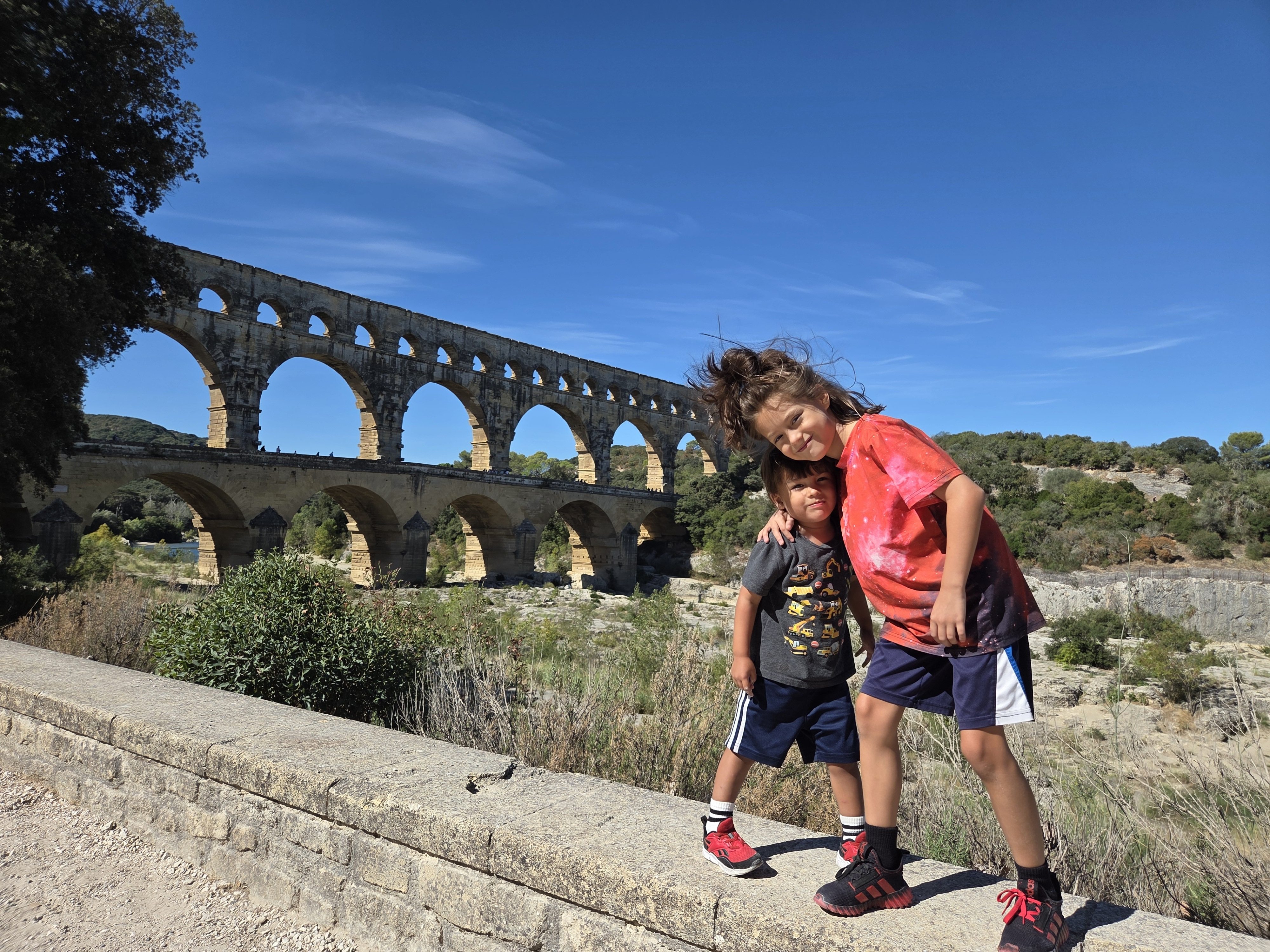 Remi and Arlo posing together on a stone wall with the Pont du Gard's three tiers of arches stretching across the background
