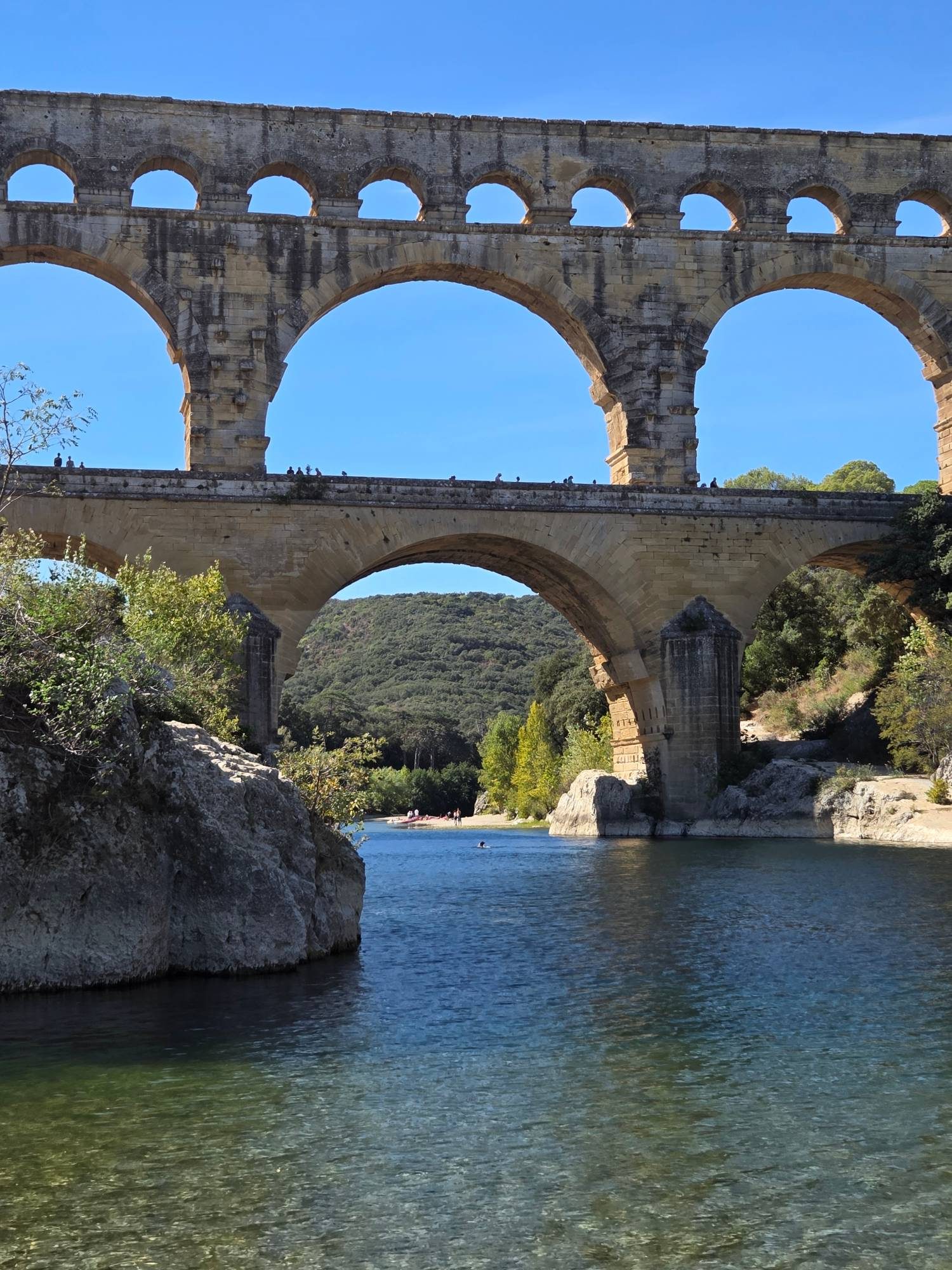 The Pont du Gard rising above the clear green-blue Gardon river from the rocky shore, a tiny swimmer in the water near the base of the bridge and people walking across the lower tier
