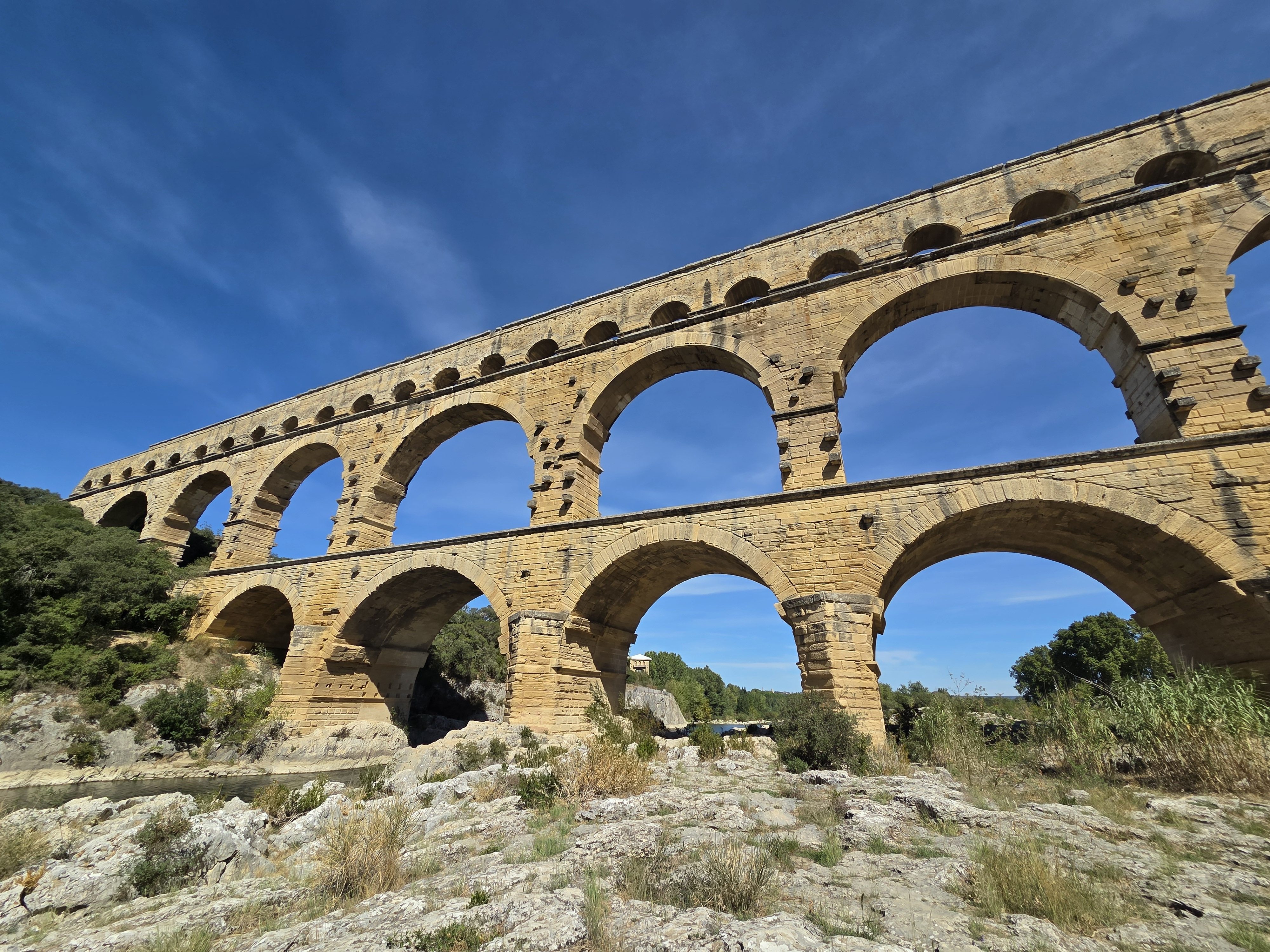 The Pont du Gard aqueduct rising in three tiers of honey-colored stone arches against a blue sky, seen from the rocky ground below
