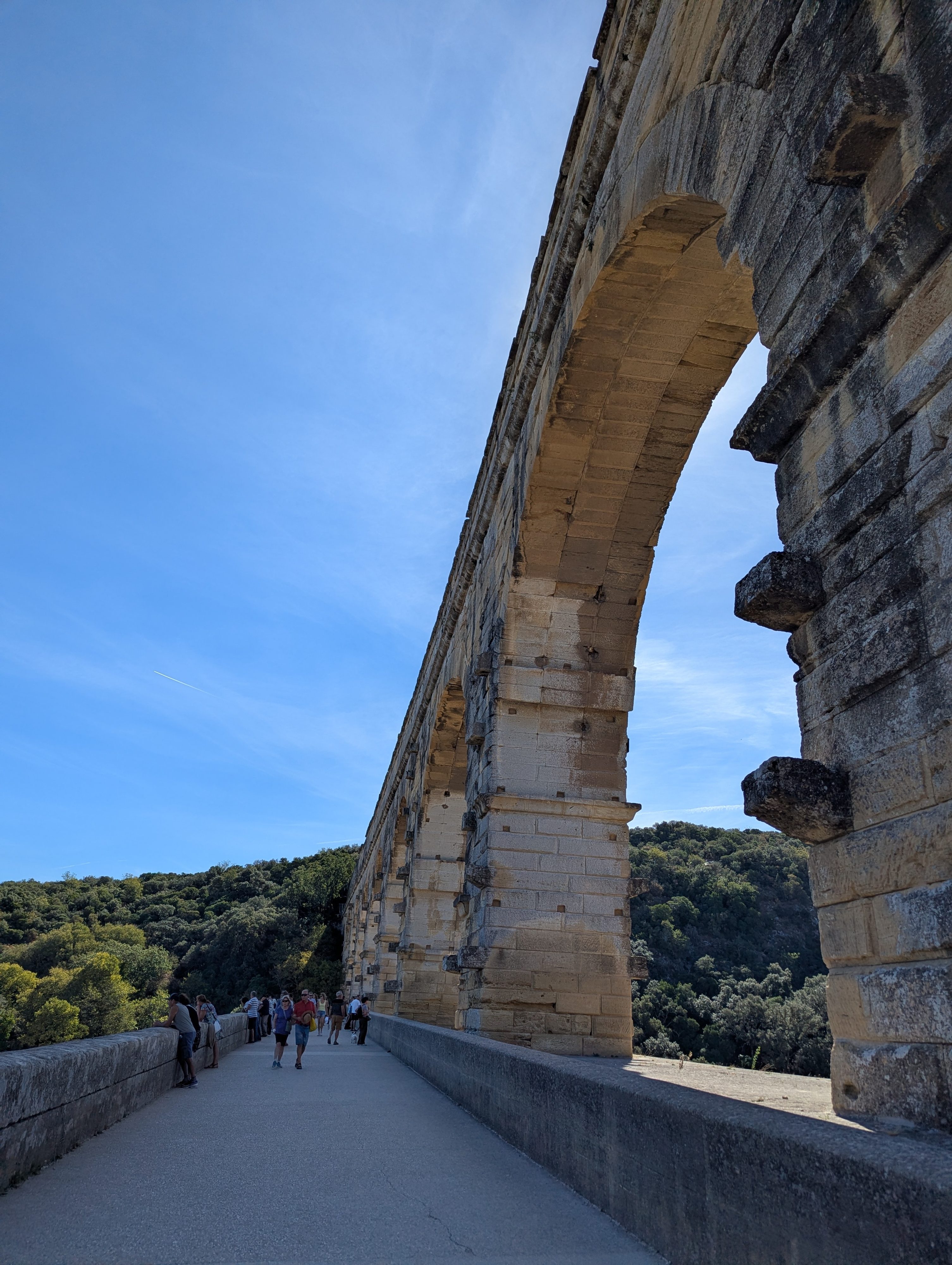 The walkway across the lower level of the Pont du Gard, the massive stone arches rising overhead with visitors walking along the path, green hills in the background