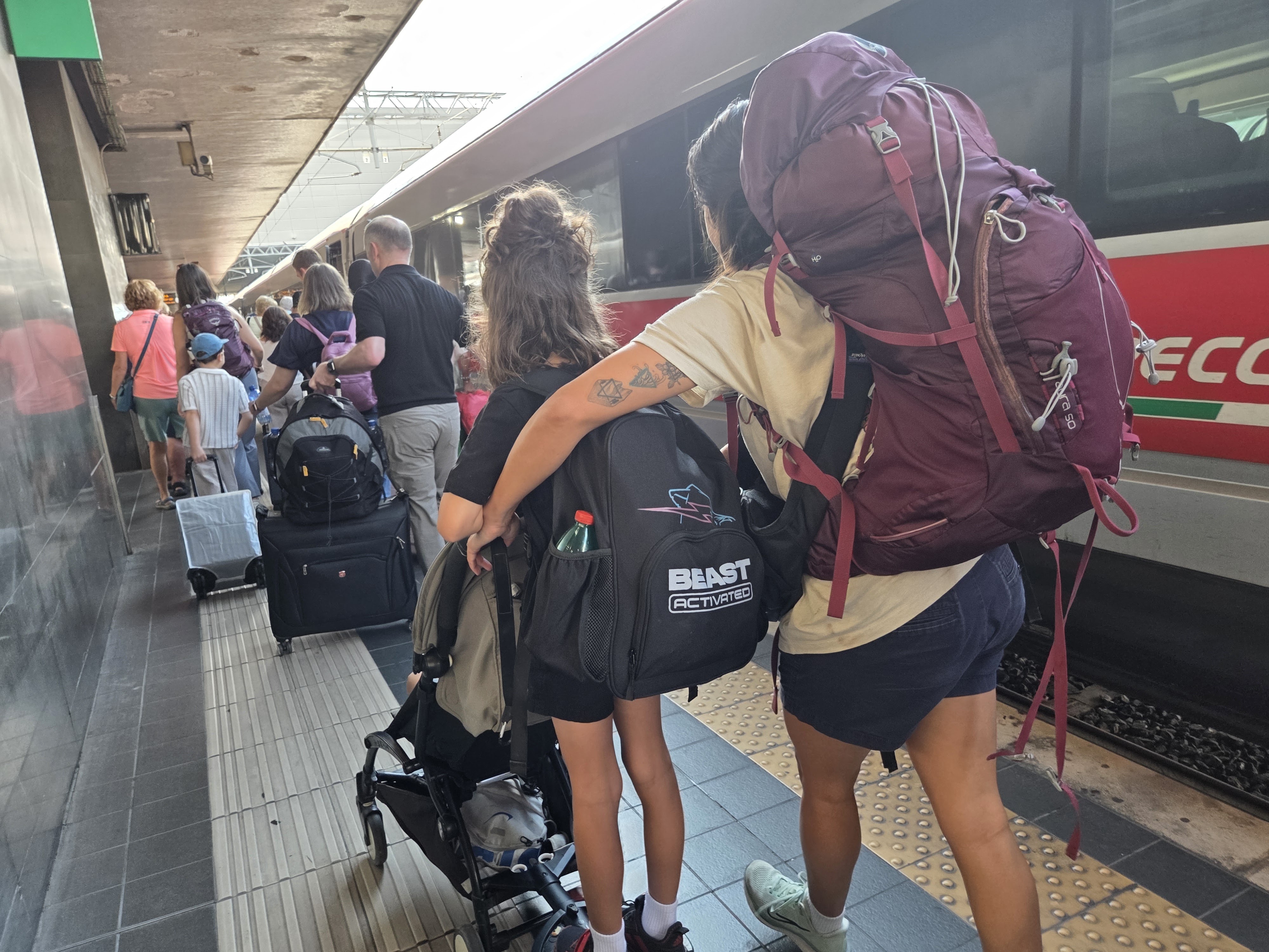 Alissa and Dani walking down a Rome Termini platform with full backpacks and a stroller, following other travelers toward a train