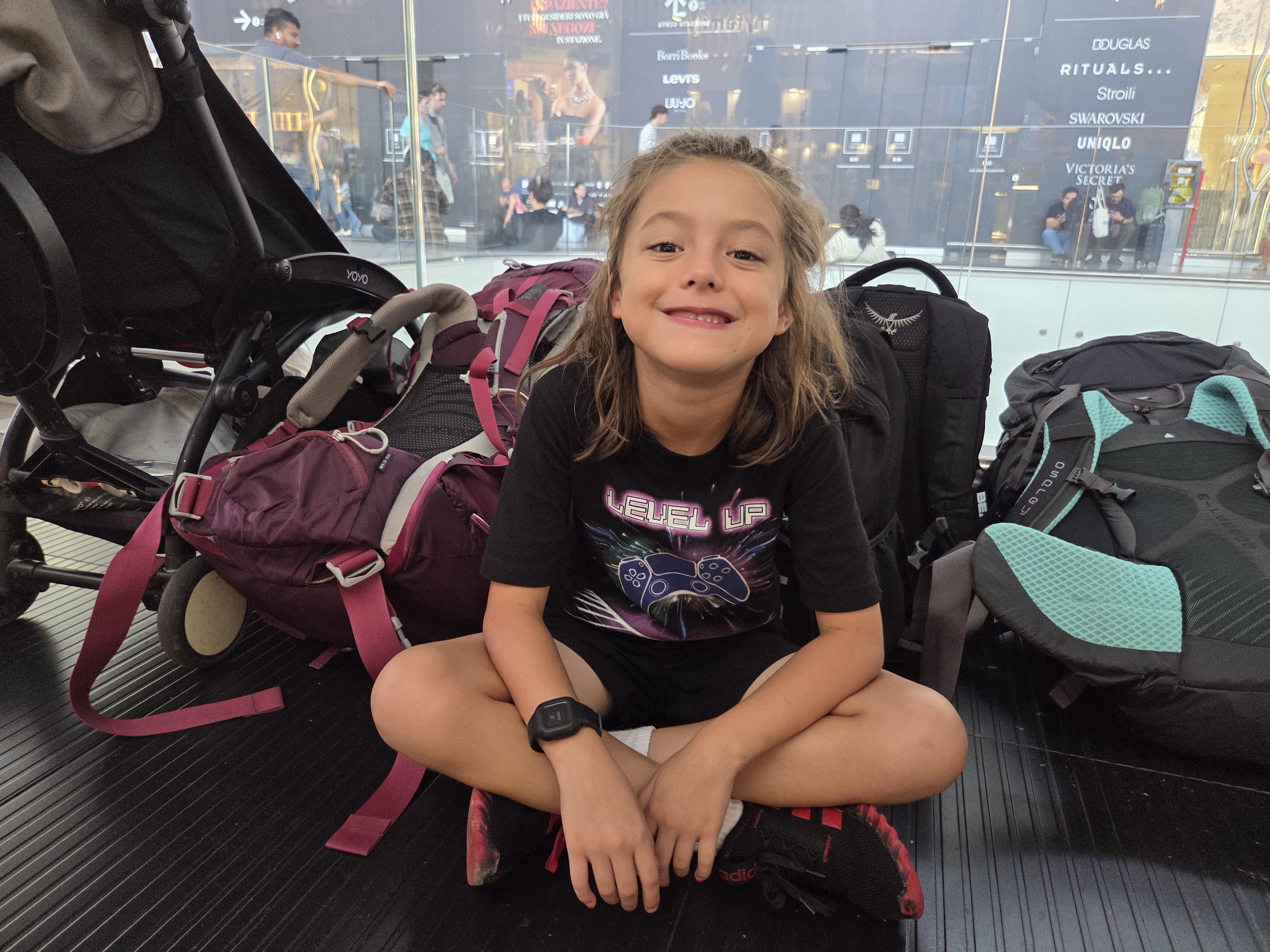 Remi sitting cross-legged and smiling on the floor of a train station beside a stroller and backpacks