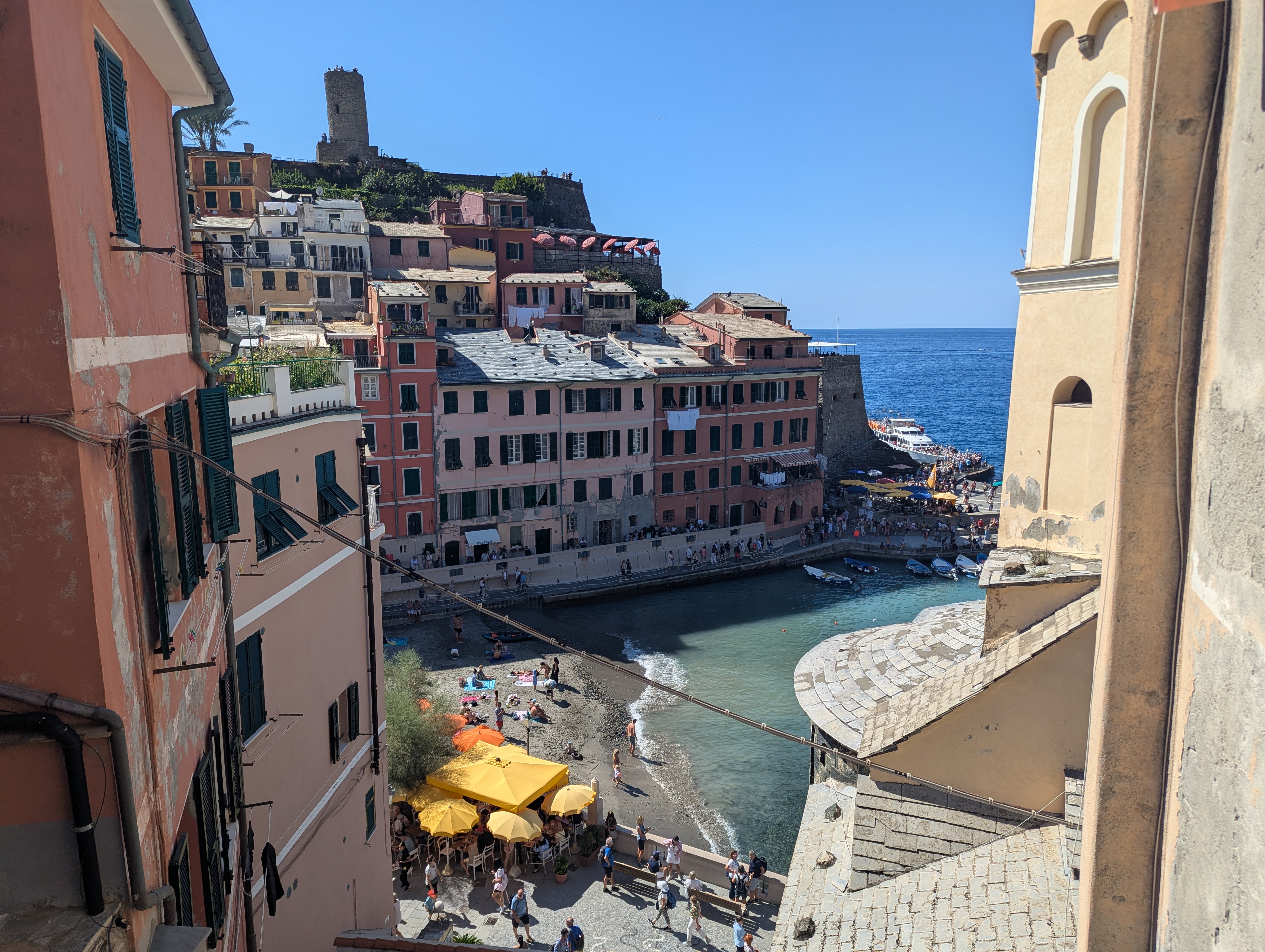 A high view down over Vernazza's harbor, pastel buildings, a beach with umbrellas, and a stone watchtower above the water
