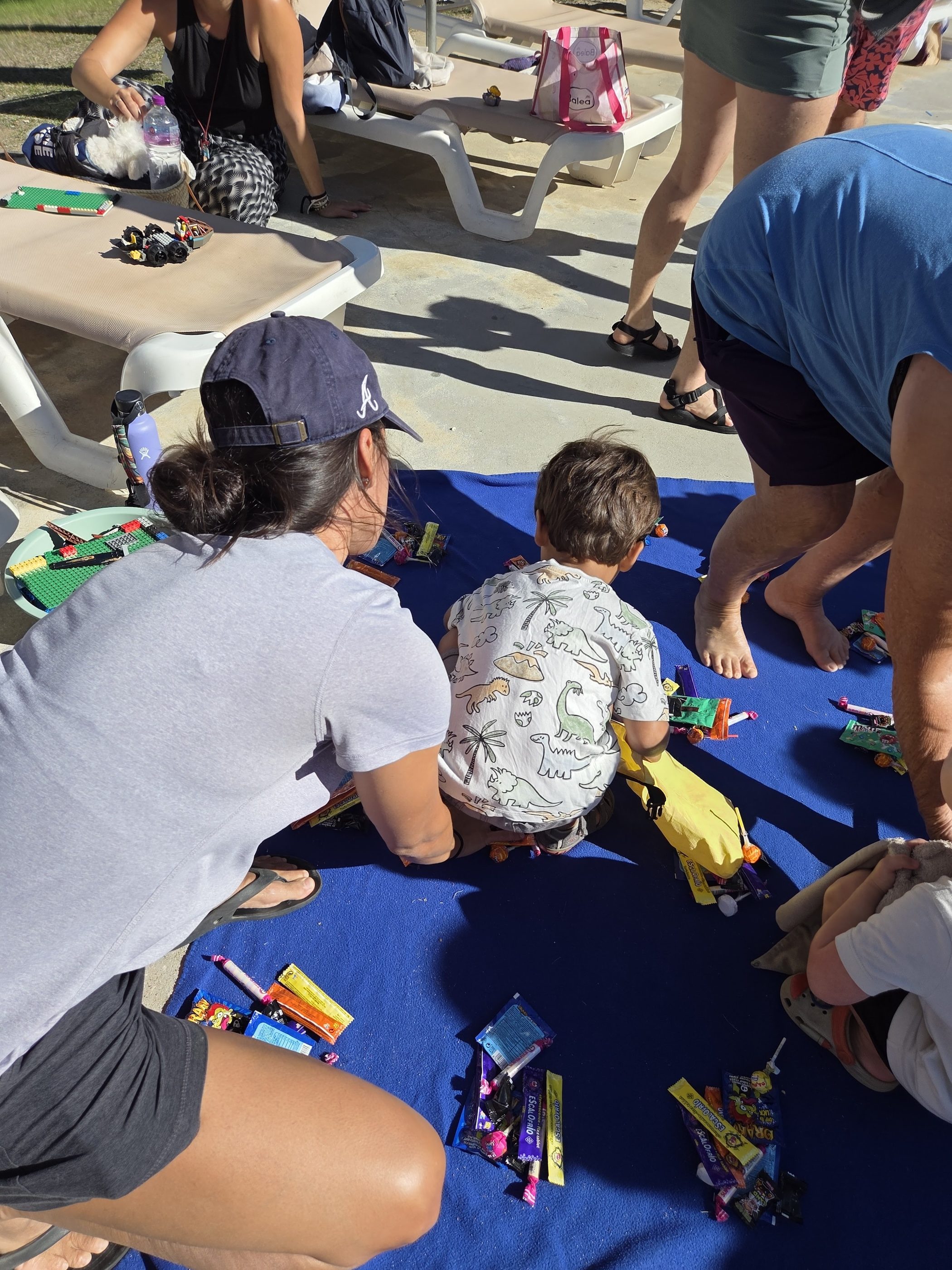 Arlo in his dinosaur T-shirt sitting on the blue blanket among the piles of candy, a parent in a baseball cap crouched beside him
