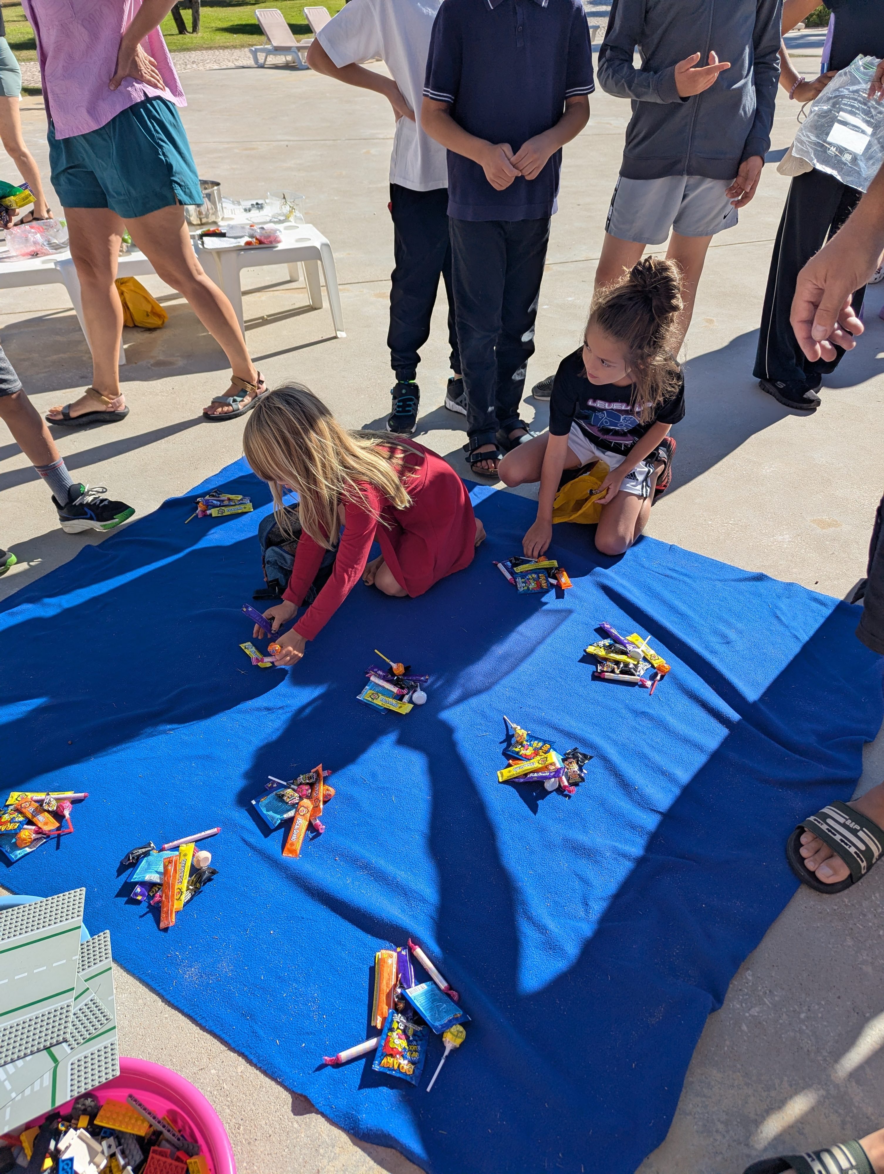 Two girls kneeling on a blue blanket by the pool, sorting candy from the hunt into even piles while other kids and parents stand around them