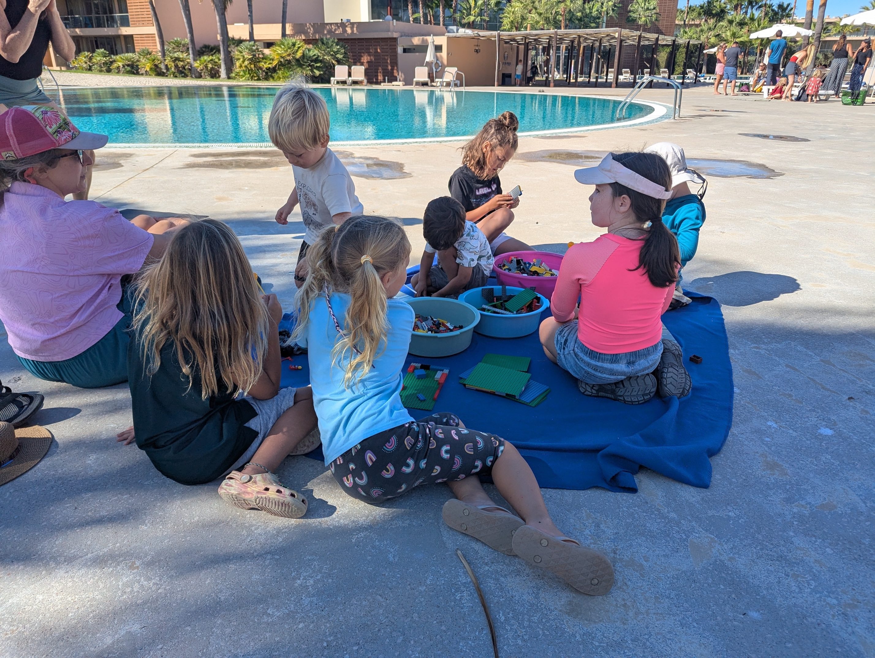 A circle of kids sitting on a blue blanket beside the pool at Herdade dos Salgados, building with plastic bricks from colorful tubs while a parent sits with them