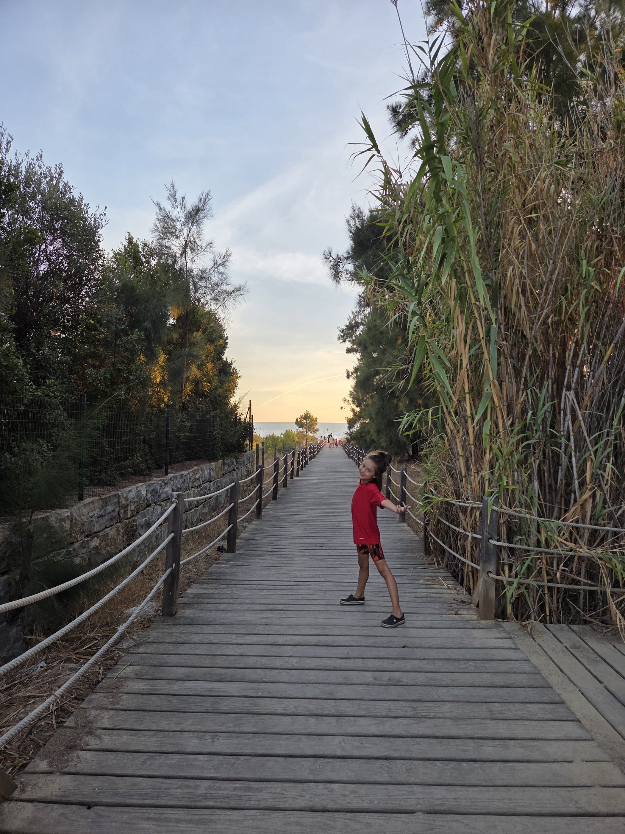 Remi in a red shirt posing on a wooden boardwalk lined with rope railings and tall reeds, the path leading to the sea under an evening sky
