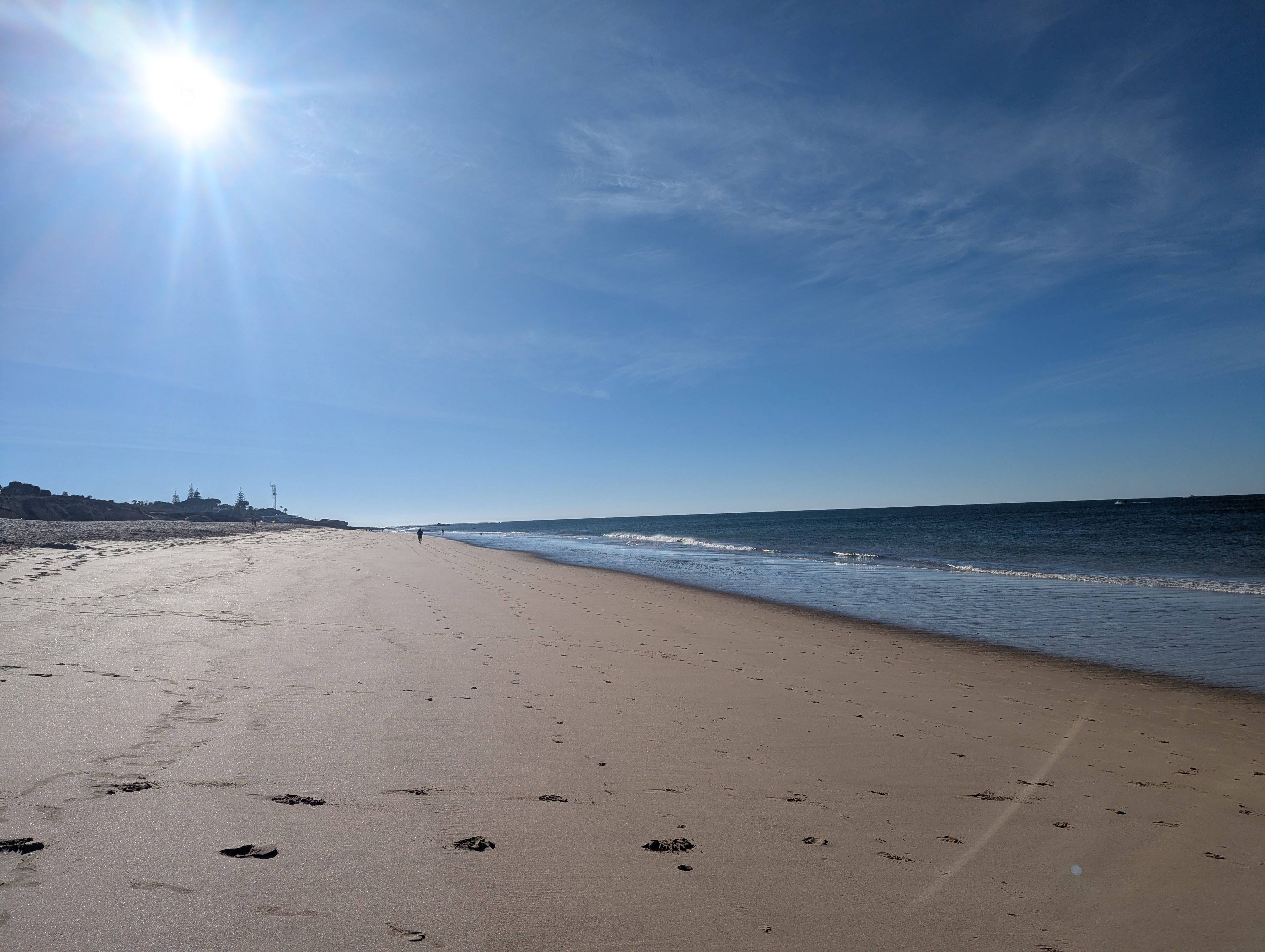A long, wide stretch of pale sand on Praia da Galé under a bright morning sun, with gentle waves on the right and a single person walking far down the beach