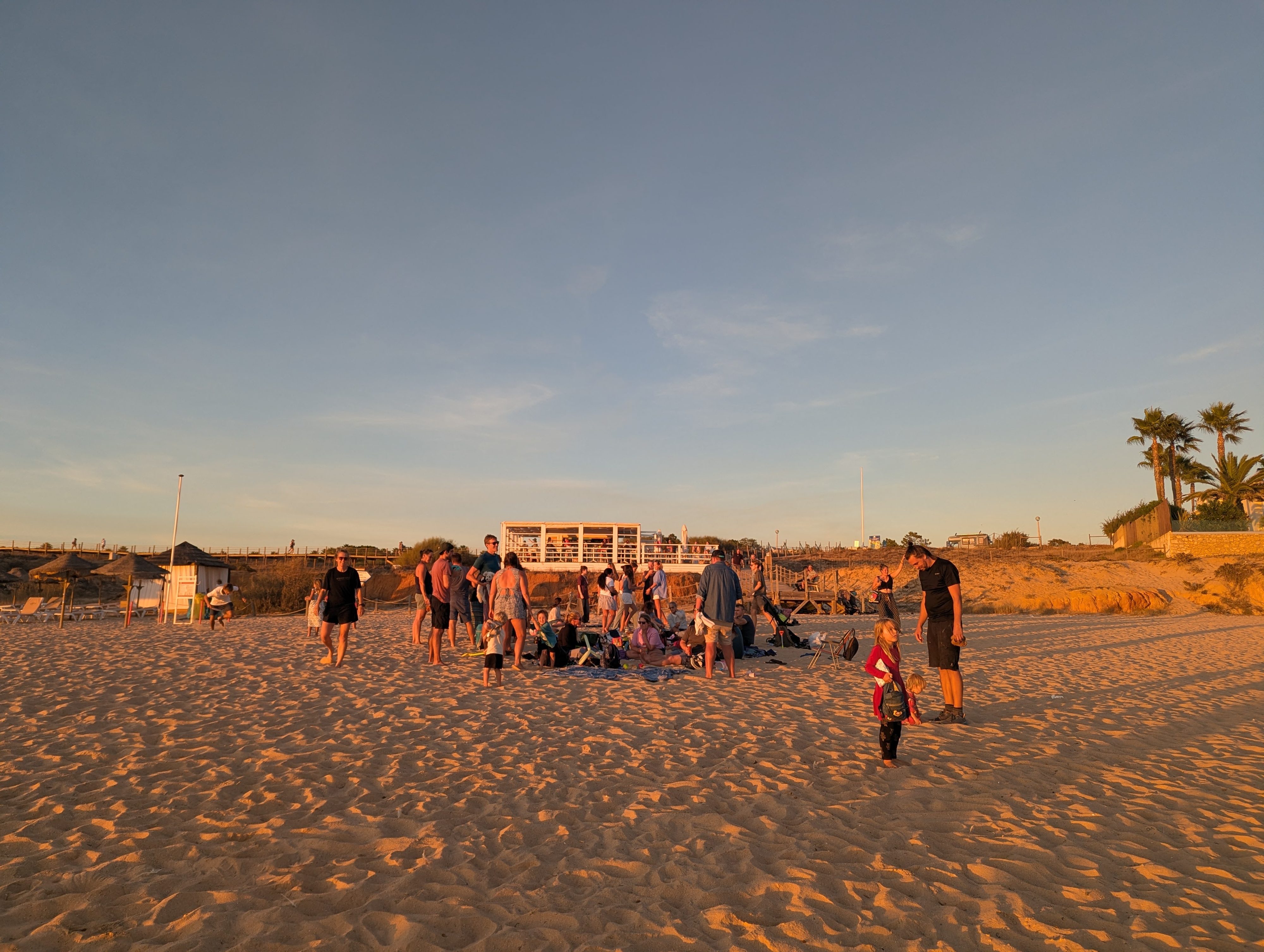 Families from the hub gathering on Praia da Galé in golden evening light, some sitting on blankets and some standing and talking, with a beach bar and palm trees behind them