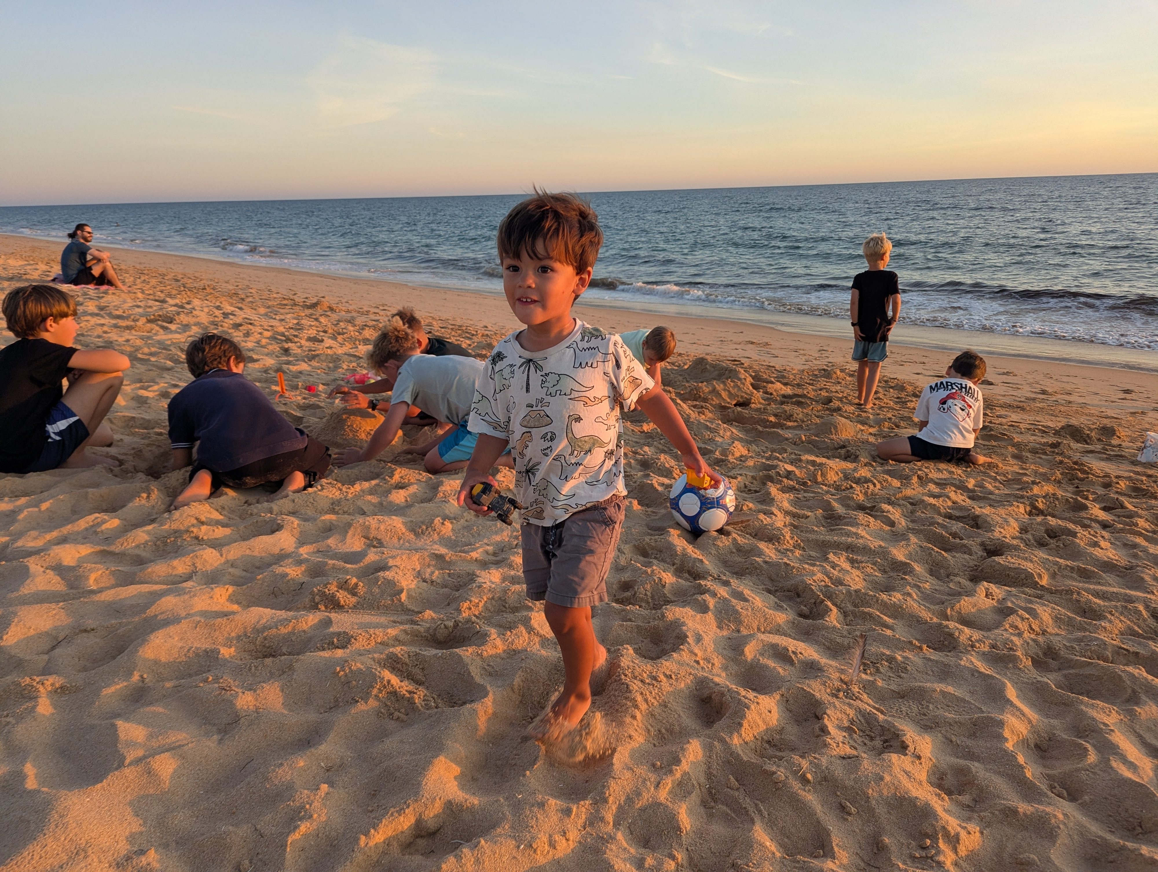 Arlo in his dinosaur T-shirt holding a soccer ball and a toy truck on the sand, with a group of kids digging behind him and the sea beyond