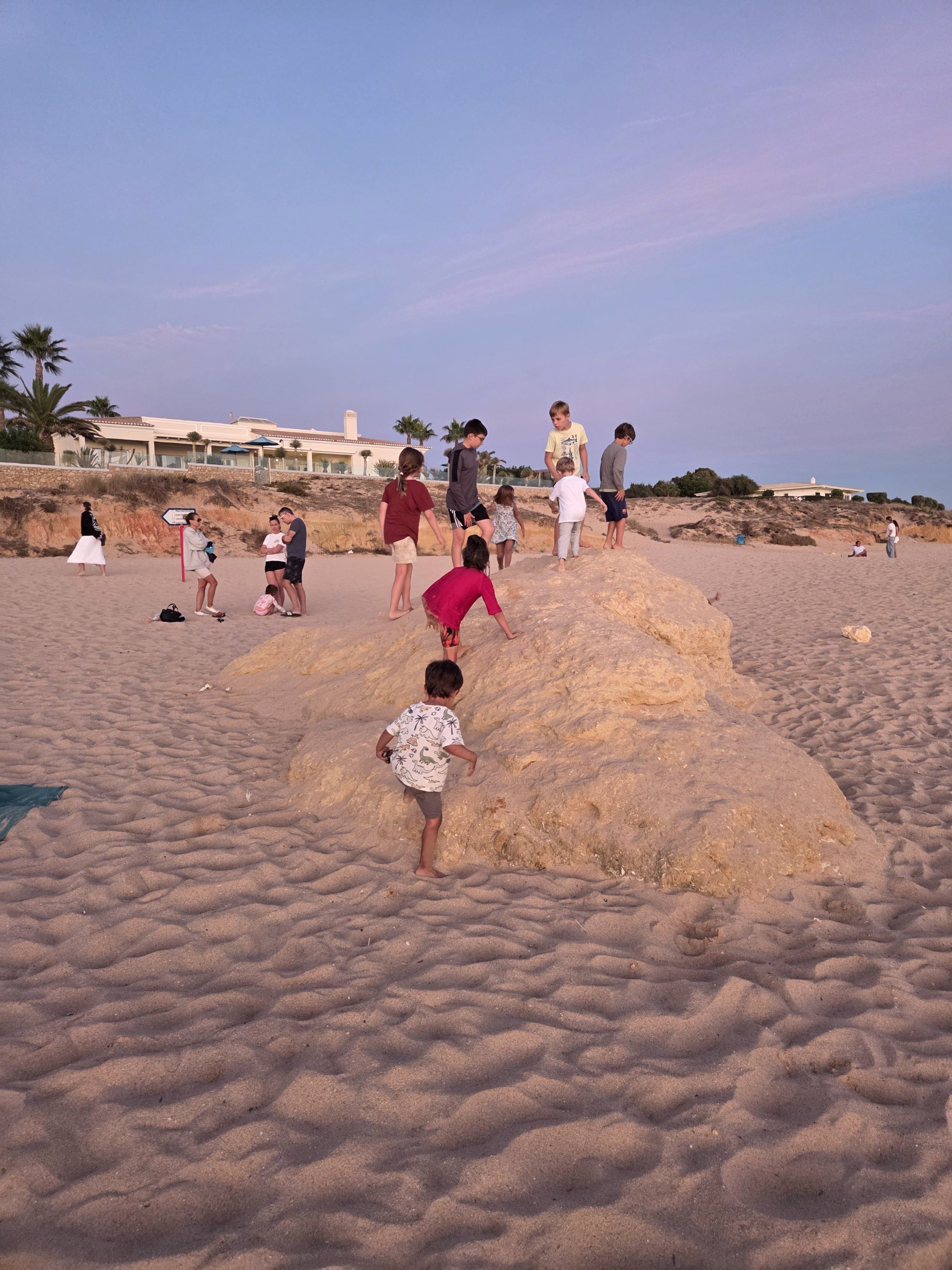 A group of kids climbing over a big sandstone rock on Praia da Galé at dusk, Arlo scrambling up the front, with parents talking on the sand and white villas and palm trees on the bluff behind