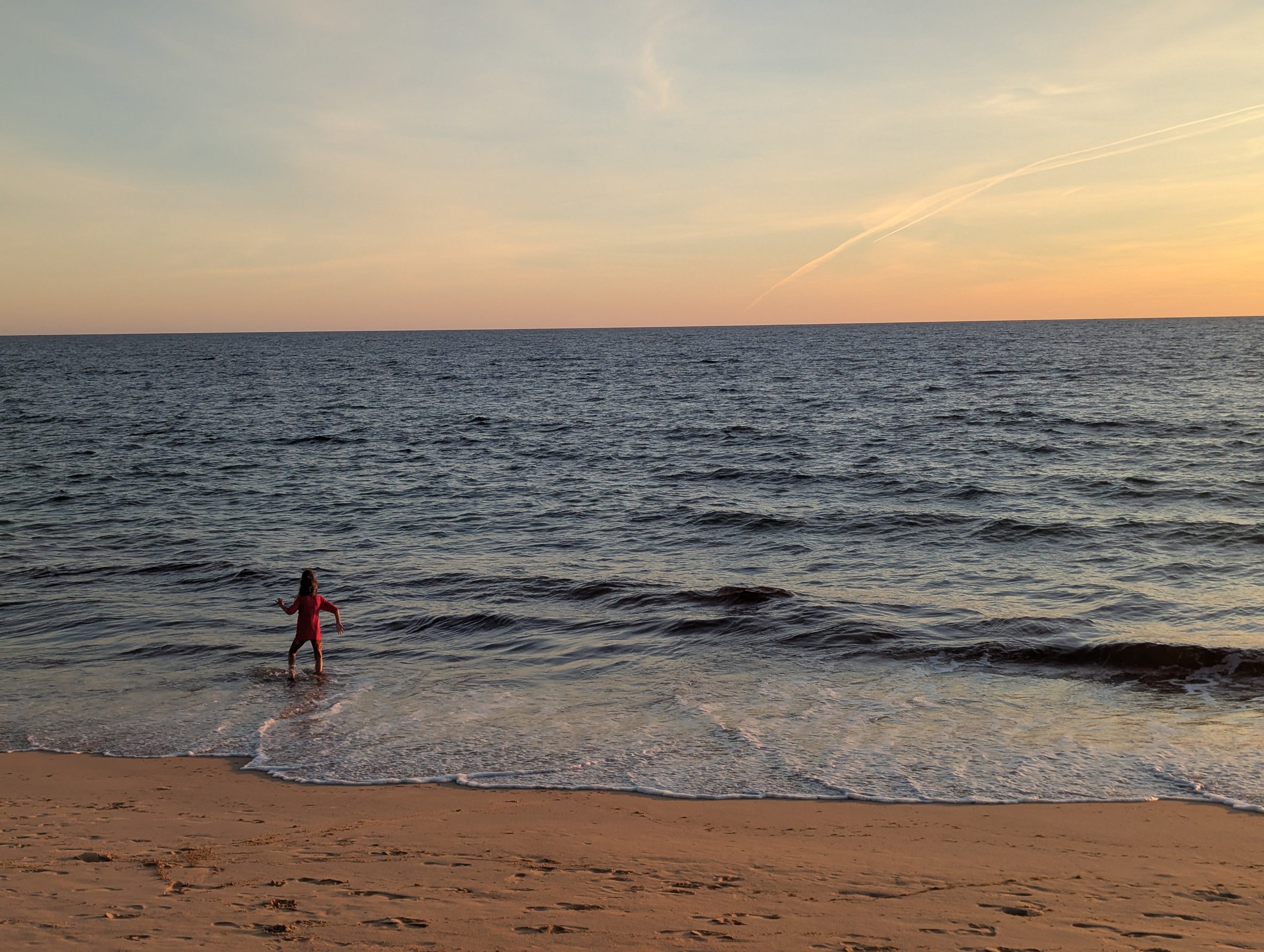 Remi in a red swimsuit wading into small waves at sunset, the sky orange and pink over the sea