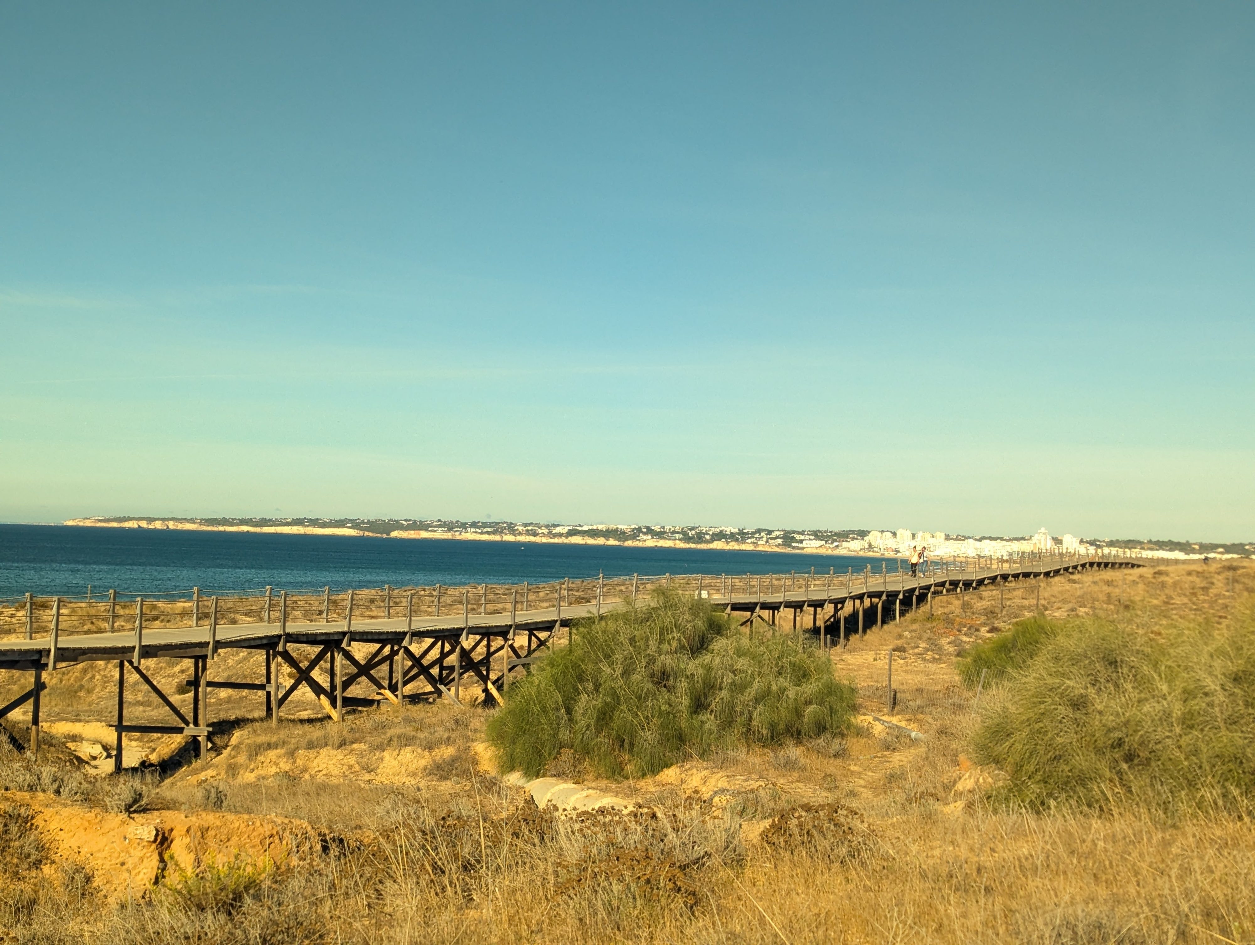 A wooden boardwalk on stilts running through dry grass and dunes along the coast, with blue sea on the left and white buildings along the shore in the distance