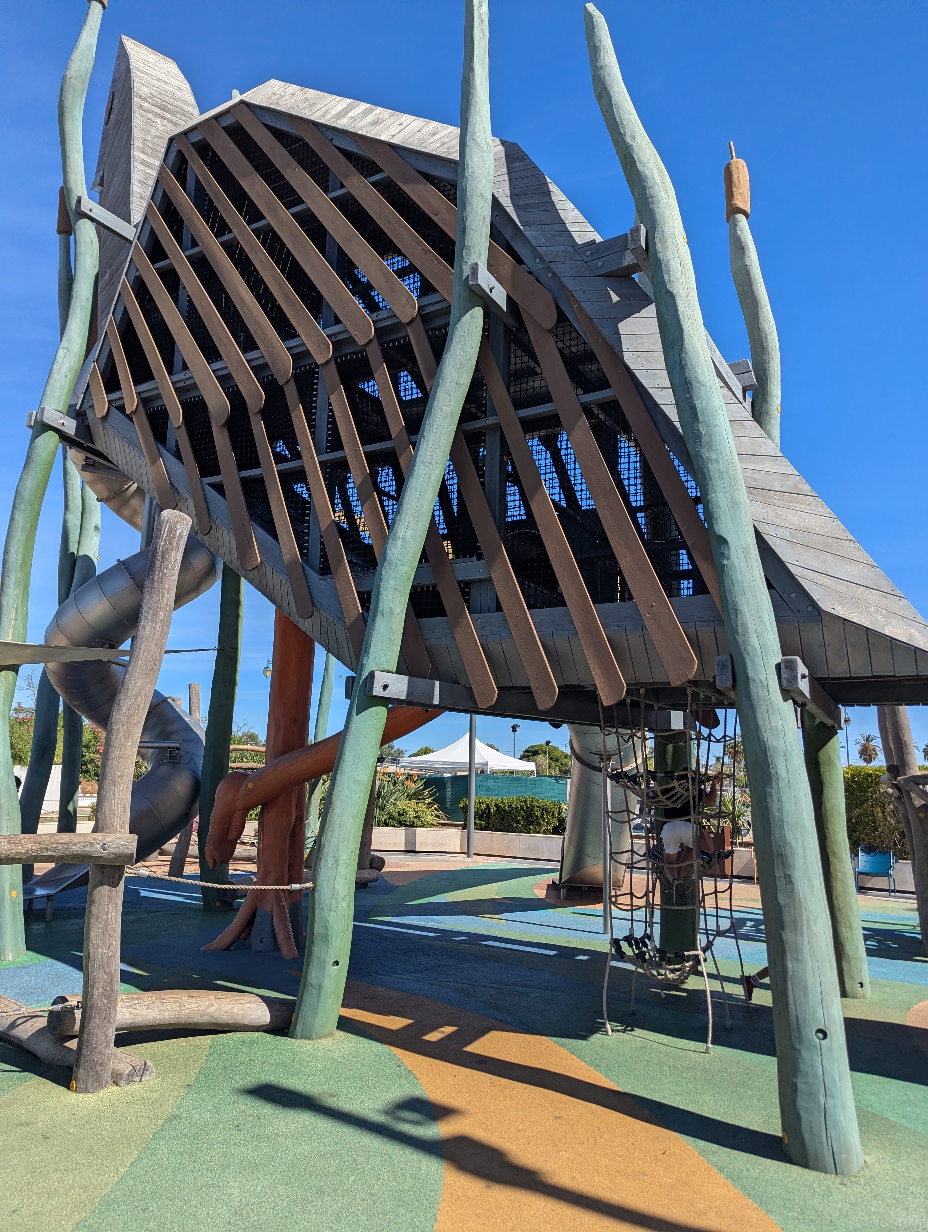 A giant wooden bird-shaped climbing structure on tall green poles at the AlgarveShopping playground, with a rope net, a silver tube slide, and colorful soft flooring under a blue sky