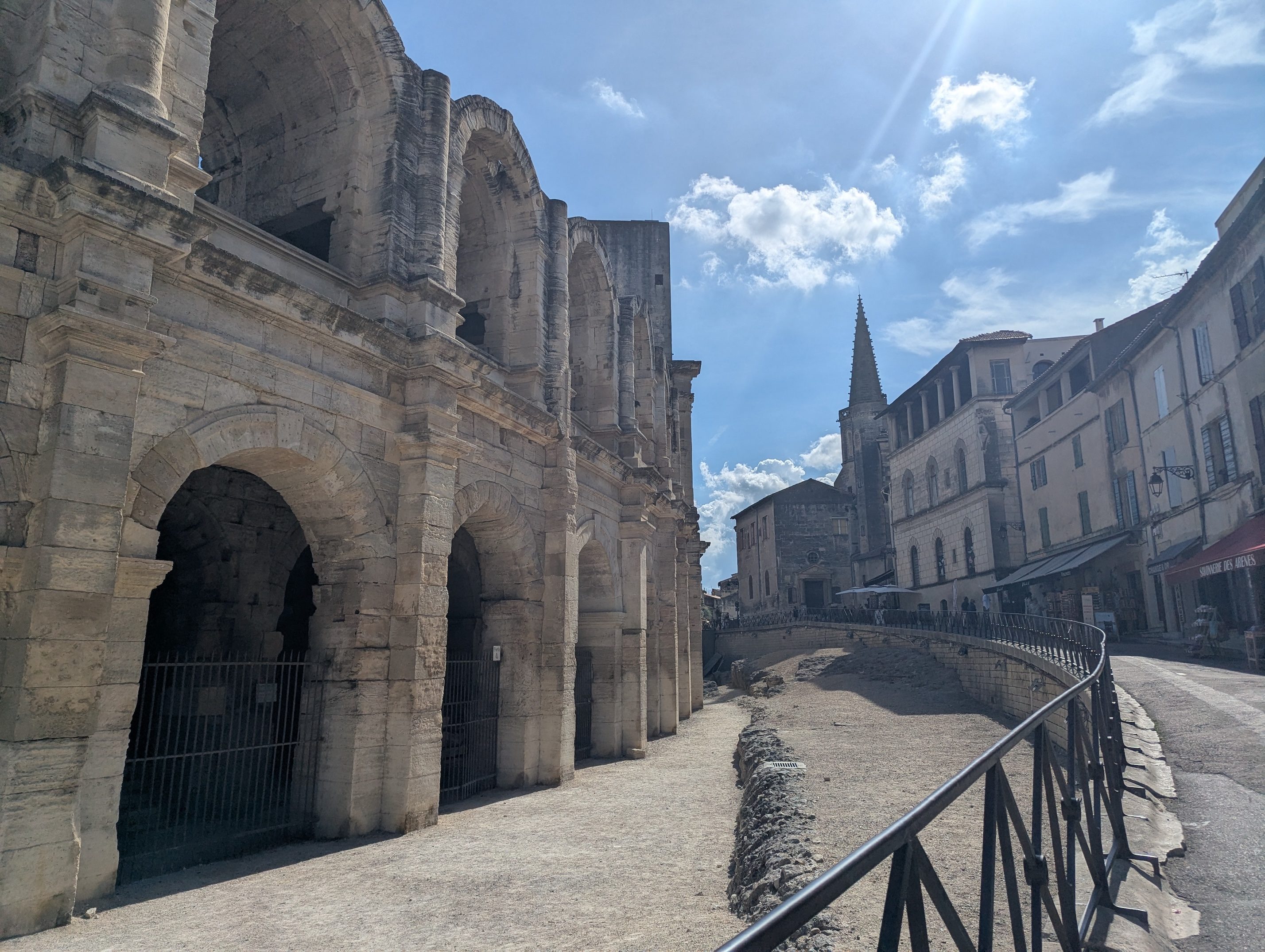 The weathered arched stone exterior of the Roman arena in Arles curving along a street, with a church steeple and old buildings beyond