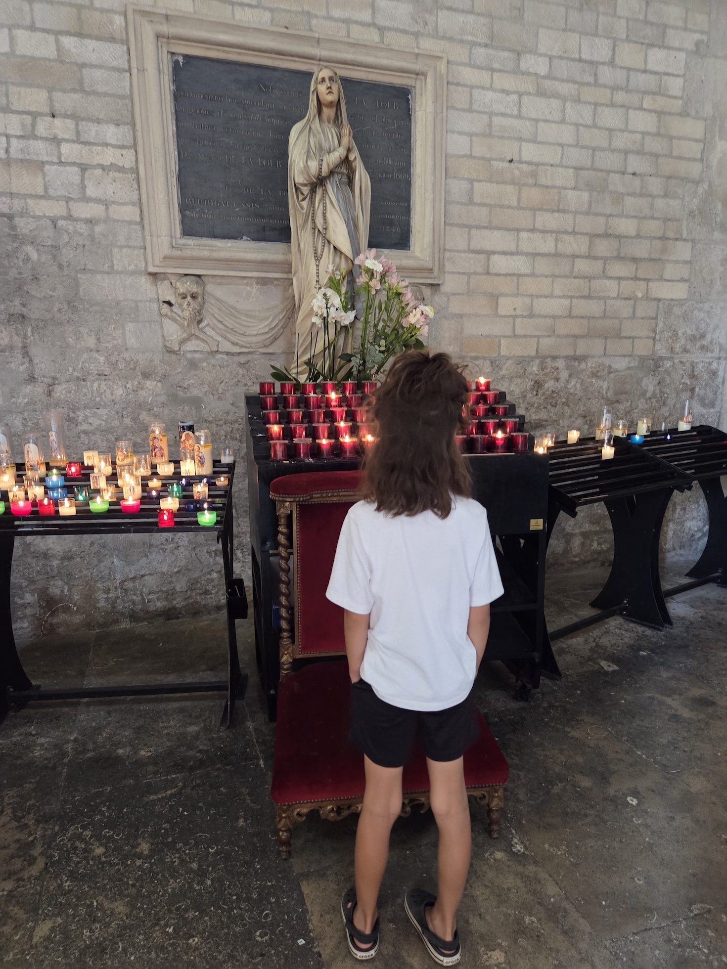 Remi standing with her back to the camera in front of a statue of the Virgin Mary and rows of red and colored votive candles in the stone church of Saint-Trophime
