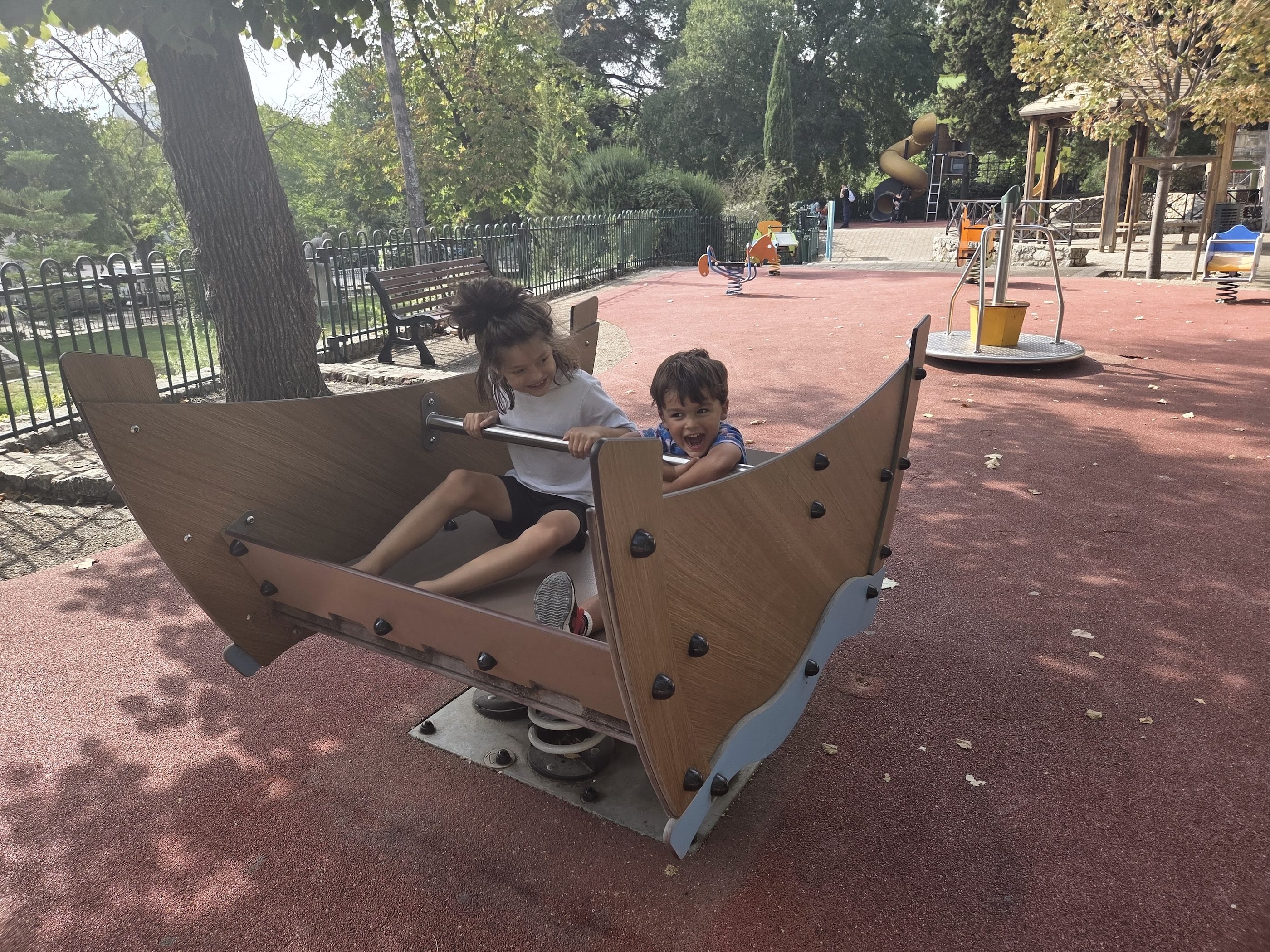 Remi and Arlo rocking together in a big wooden boat-shaped spring rocker on a red rubber playground surface under tall trees