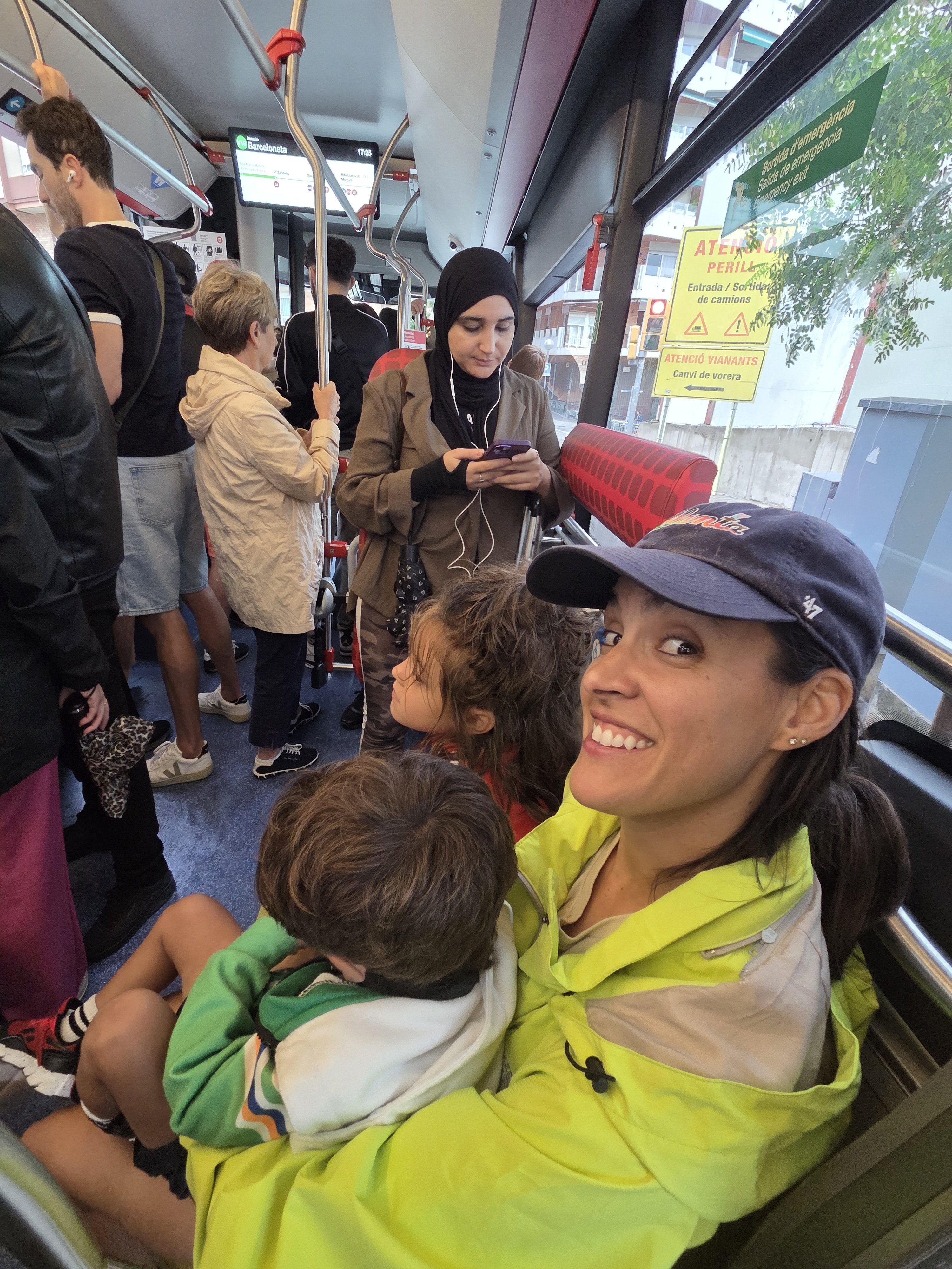 Alissa in a cap and lime-green rain jacket smiling at the camera on a crowded city bus, Arlo curled in her lap and Remi beside her, with a screen above showing Barceloneta as the destination