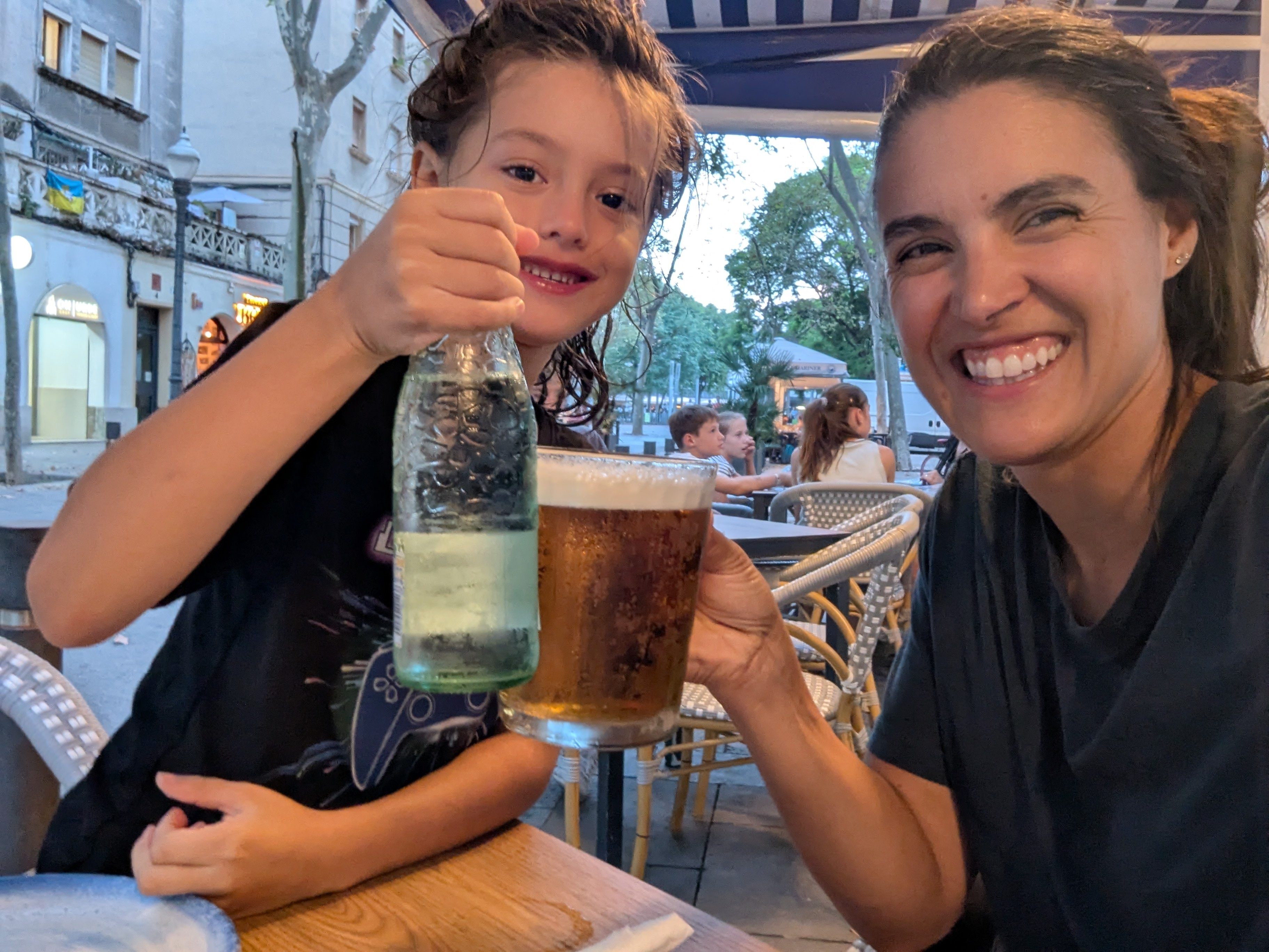 Remi holding up a glass bottle and Alissa holding a pint of beer, clinking them together at a sidewalk cafe table at dusk, both grinning