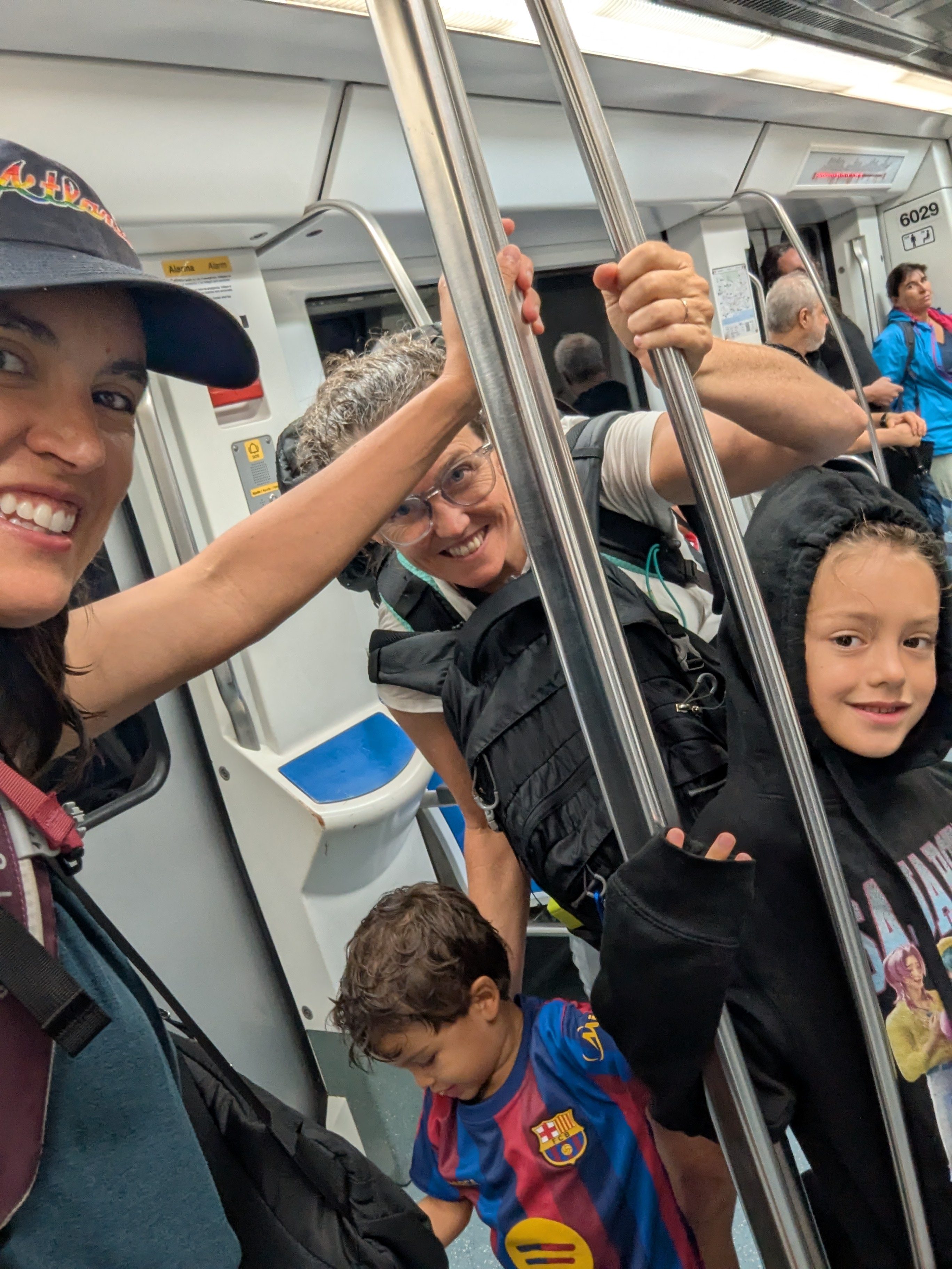 Alissa, Dani, Remi and Arlo squeezed into a crowded Barcelona metro car holding the poles, everyone wearing backpacks, Remi in a black hoodie and Arlo in a Barça jersey