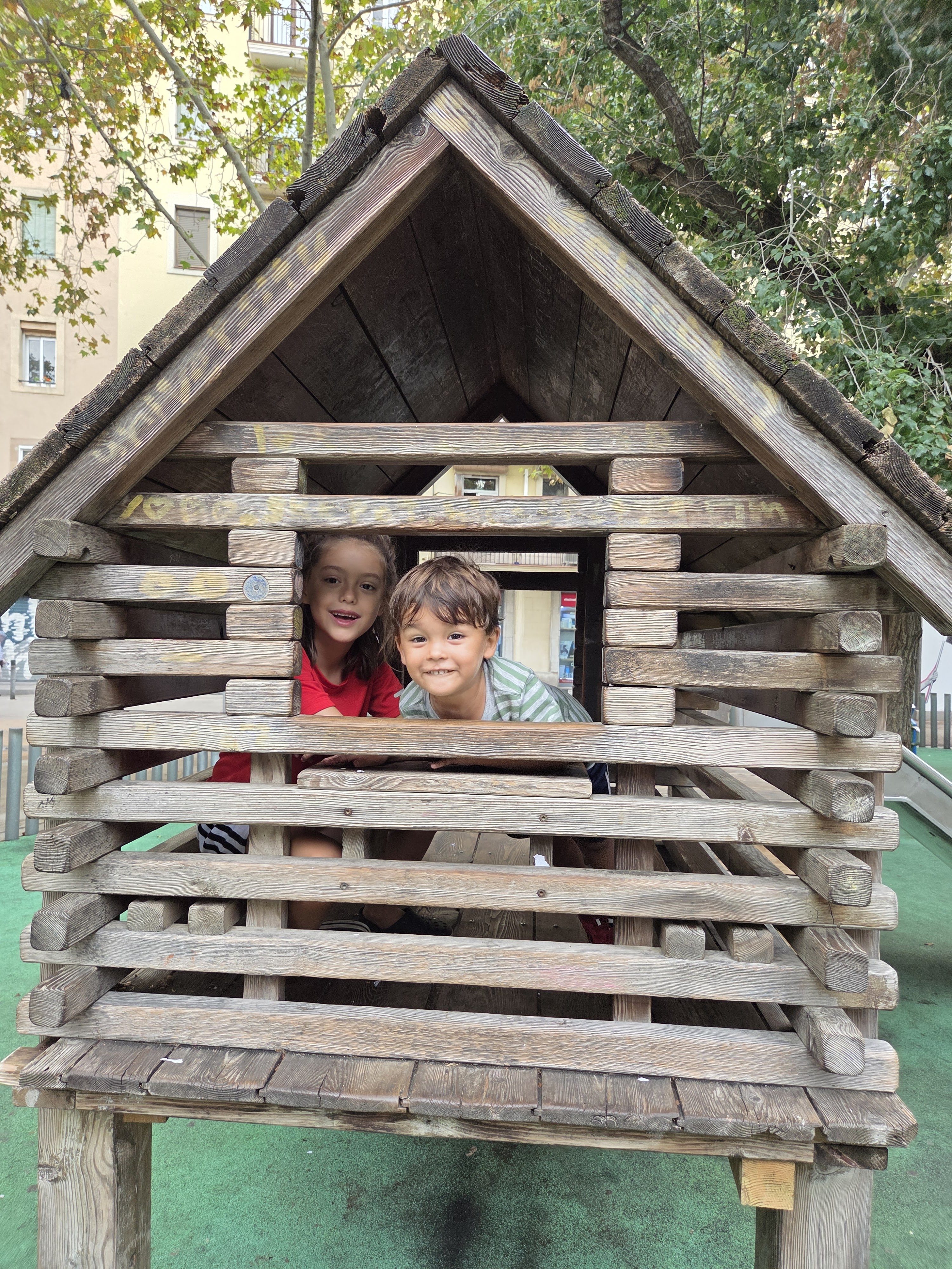 Remi and Arlo peeking out of the window of a wooden cabin on a neighborhood playground