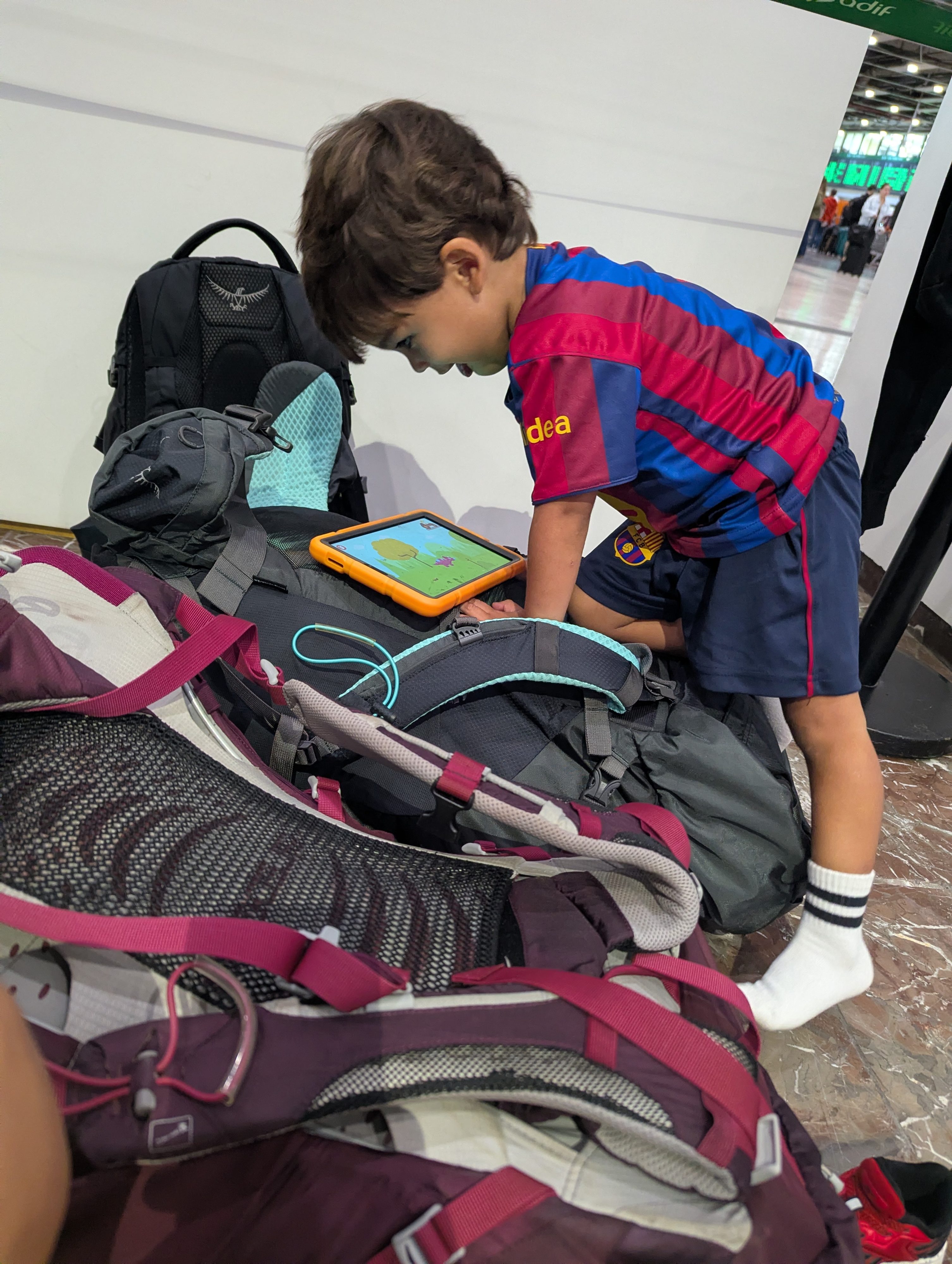 Arlo in a Barça jersey kneeling on a pile of backpacks to play on an orange-cased tablet in the train station