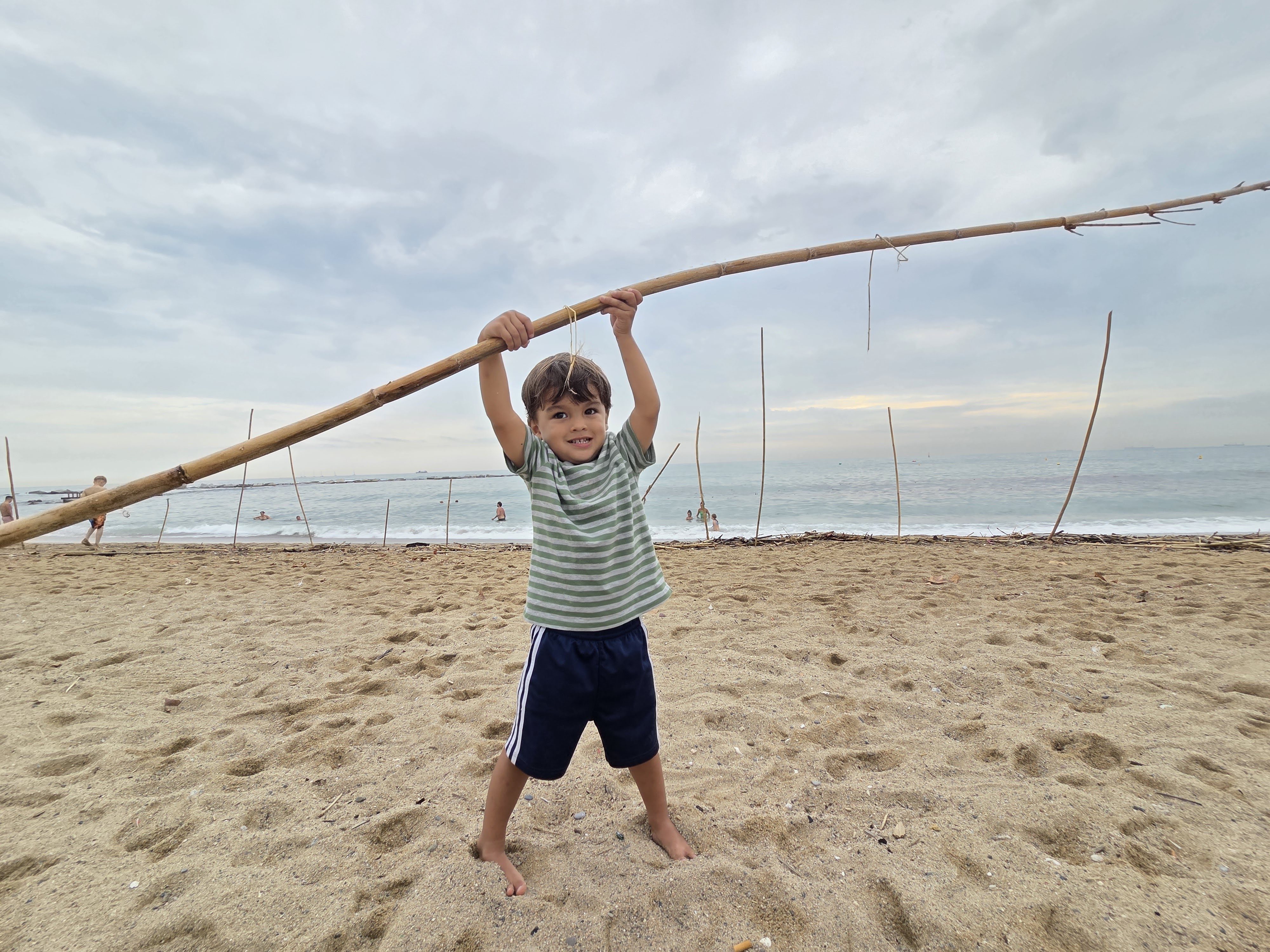 A boy standing on Barceloneta beach lifting a huge washed-up bamboo pole over his head, the sea behind him