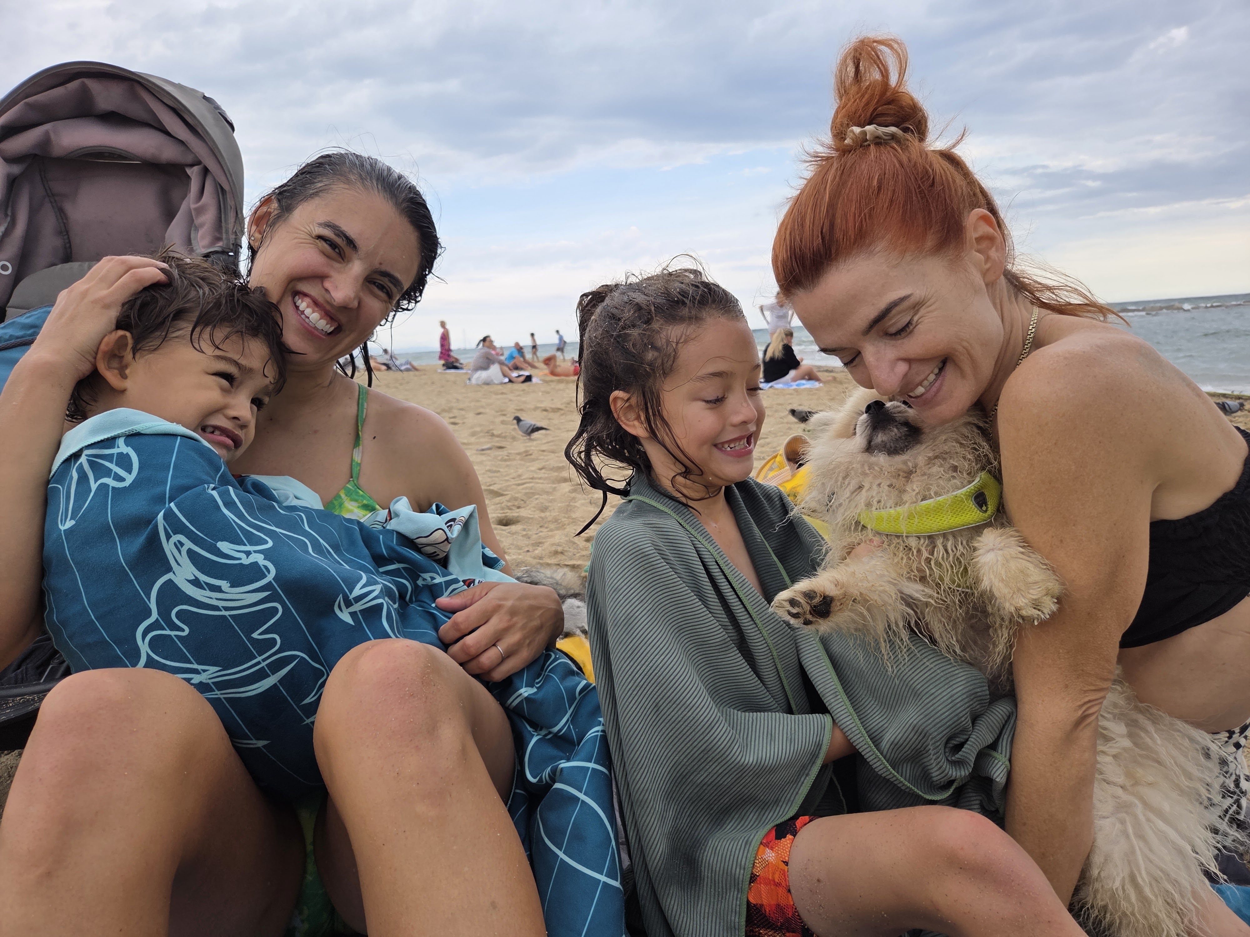 Remi, Arlo and two adults laughing on Barceloneta beach with a little fluffy dog and a stroller behind them