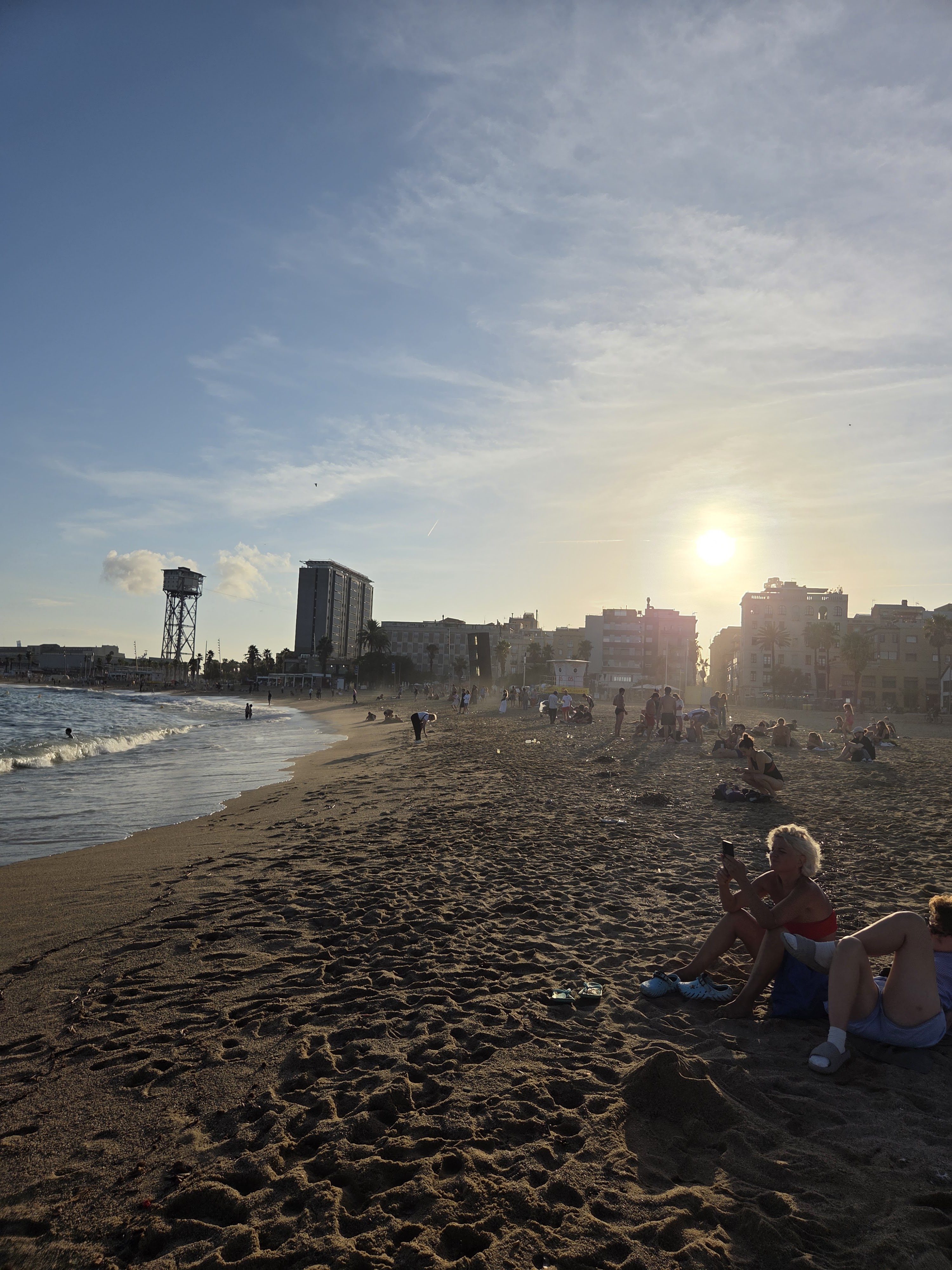 Barceloneta beach at sunset, with a cable car tower and city buildings along the shore and a few people on the sand