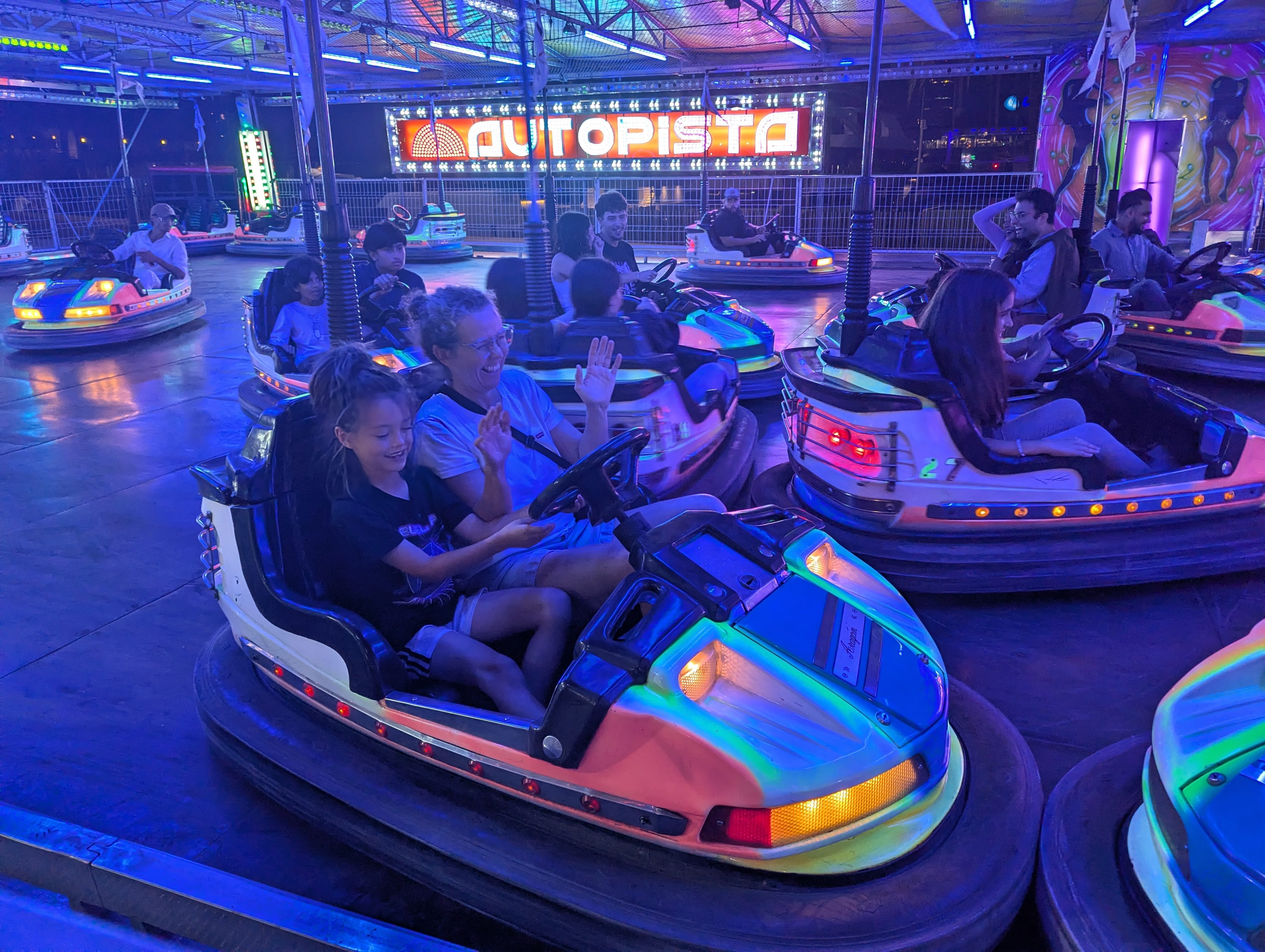 Remi and Dani laughing together in a bumper car under the colorful lights of the Autopista ride, other riders around them