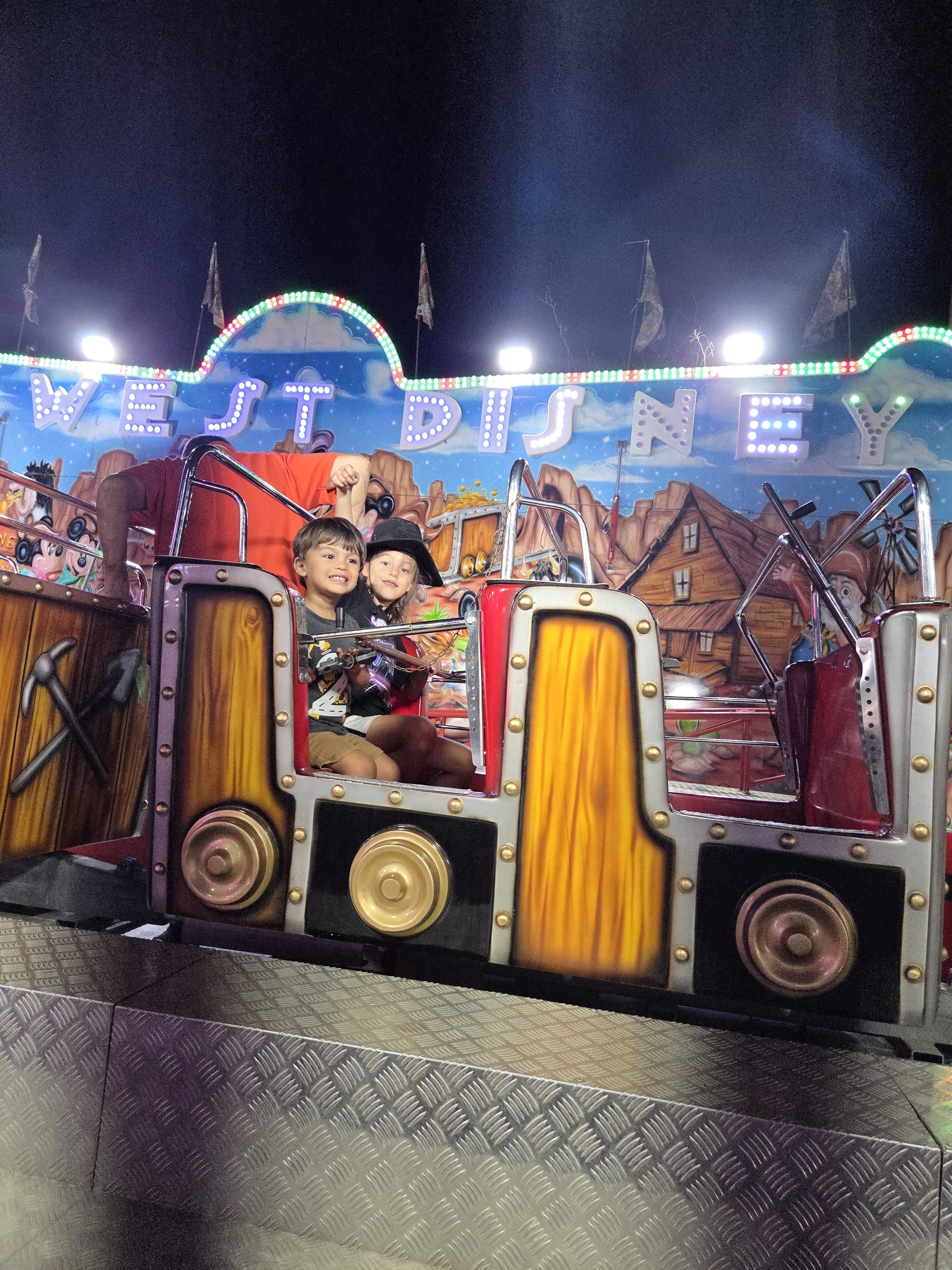 Two kids riding in a little cart on a toddler ride at night, with a colorful Wild West style facade behind them