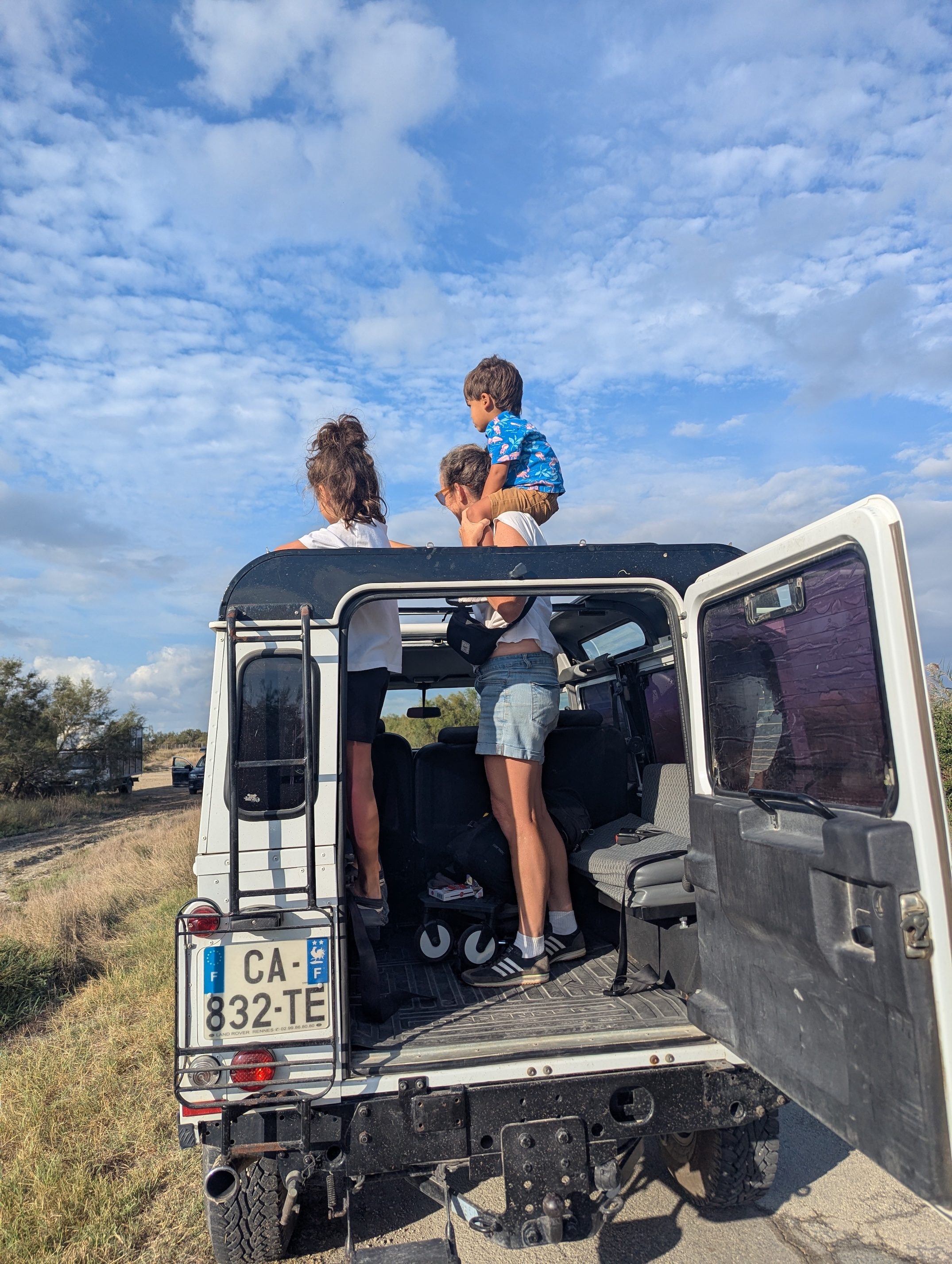 The family standing in the open back of the white safari jeep on the trail, Dani holding Arlo up, under a big blue sky