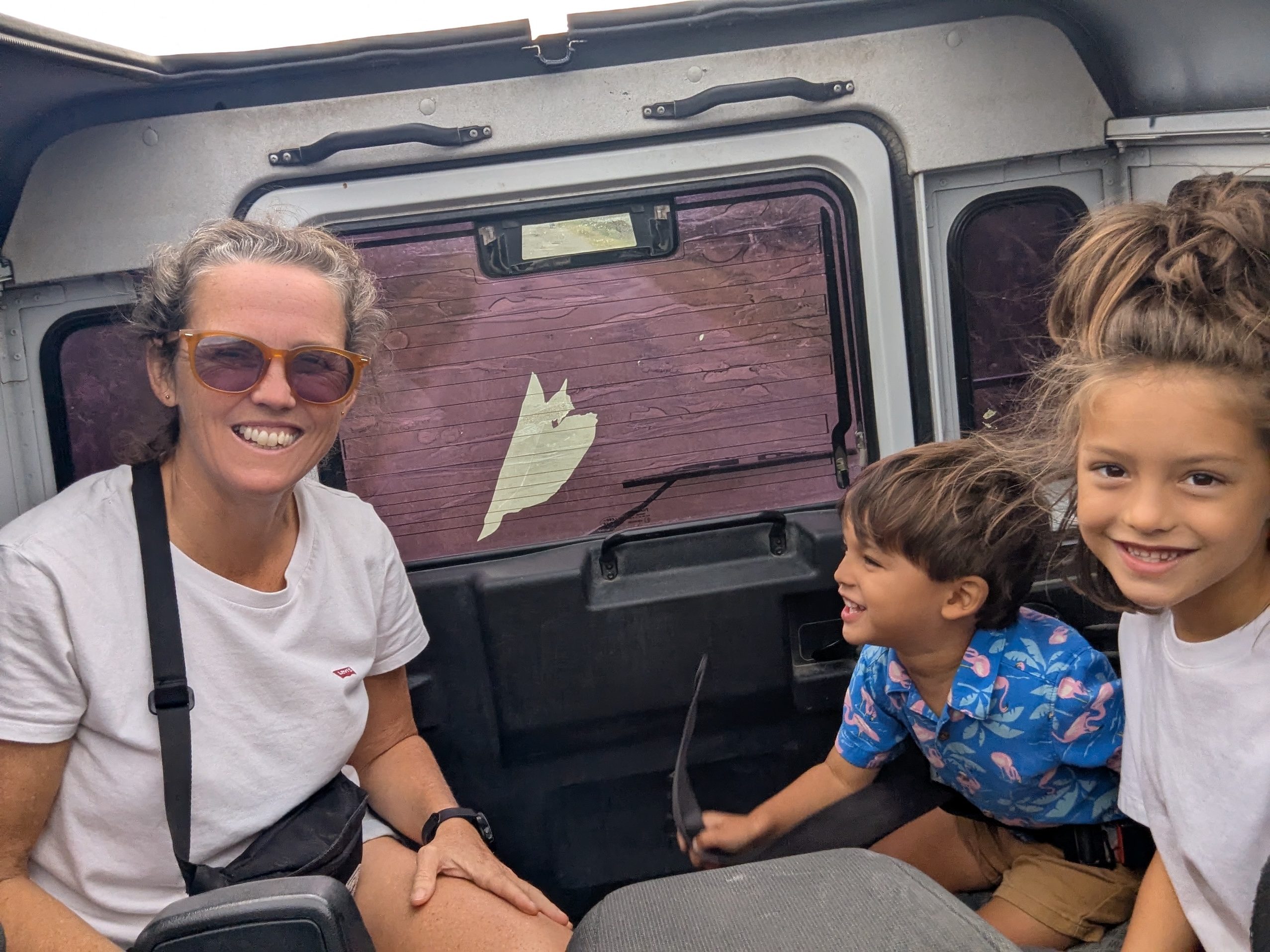 Dani smiling in sunglasses and a white shirt in the back of the safari jeep, with Arlo in a blue flamingo-print shirt and Remi grinning beside her