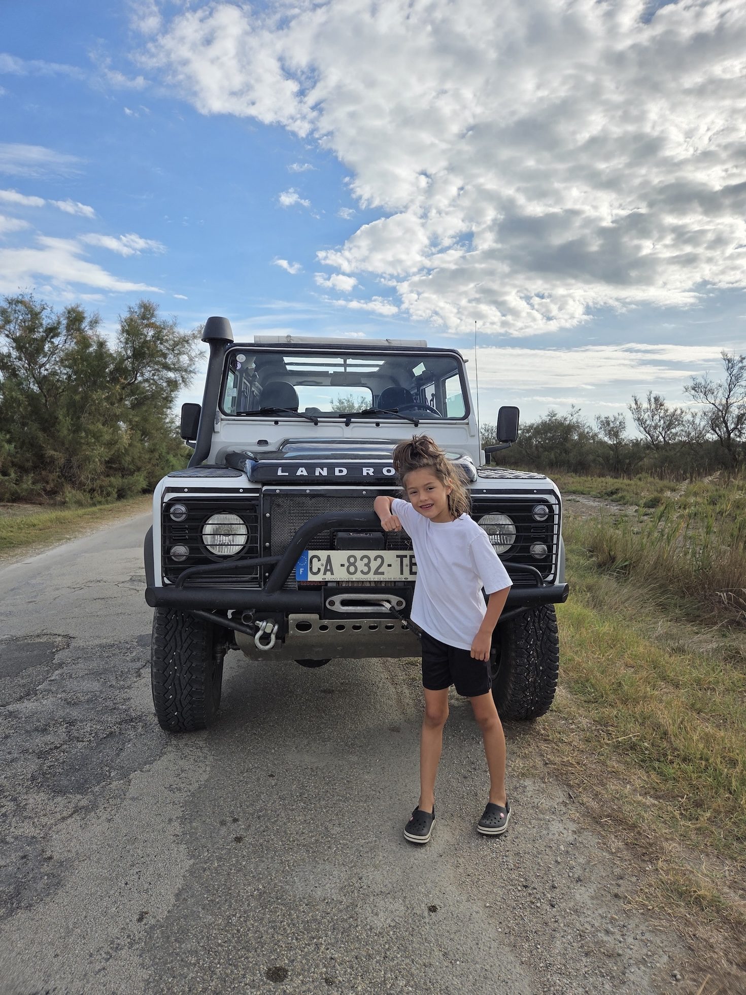 Remi leaning against the front bumper of a Land Rover safari jeep on a country road, a blue sky with big clouds behind her