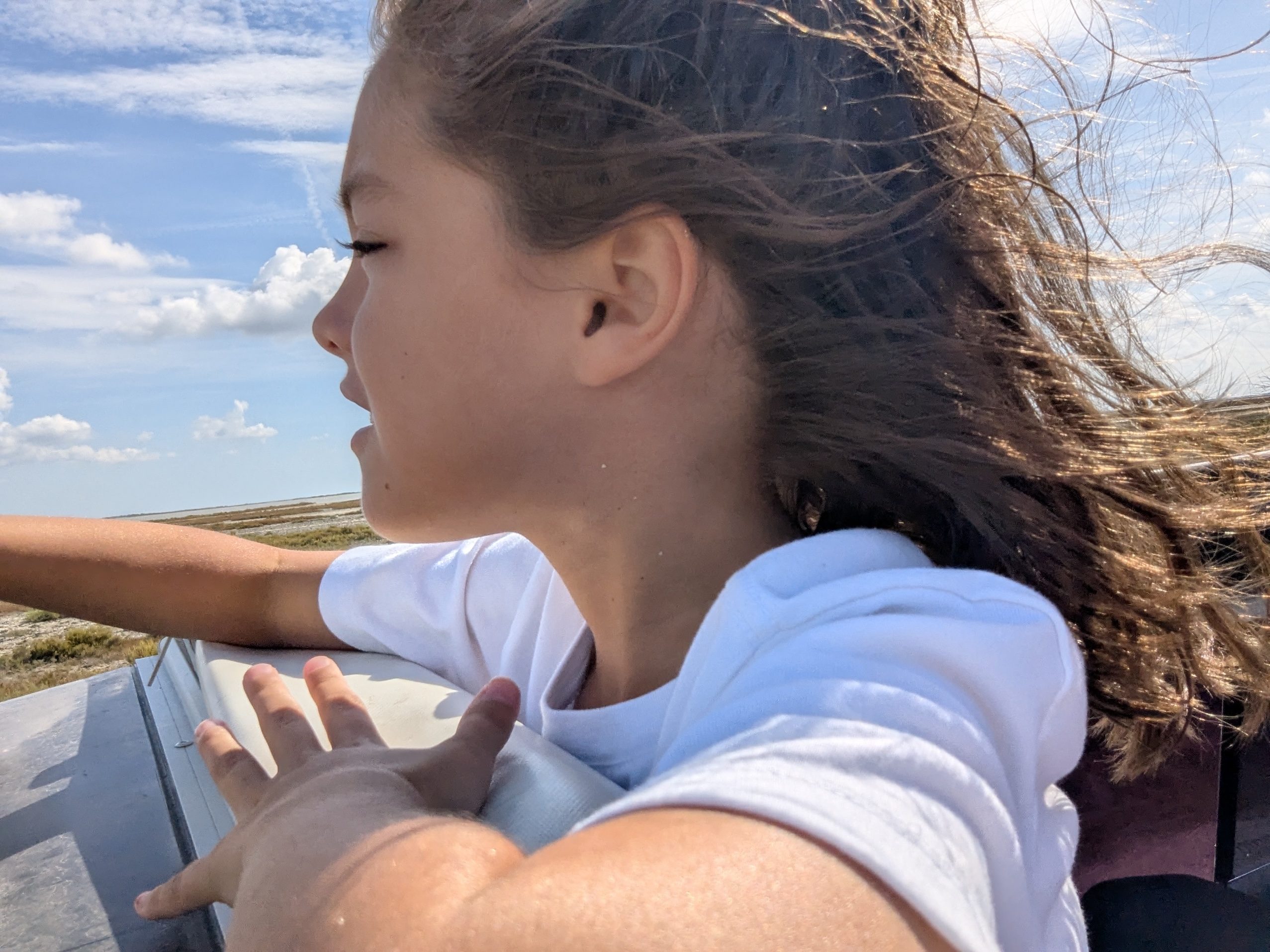 A close profile of Remi with her hair streaming back in the wind, one hand on the edge of the jeep roof, sky and marsh blurring past