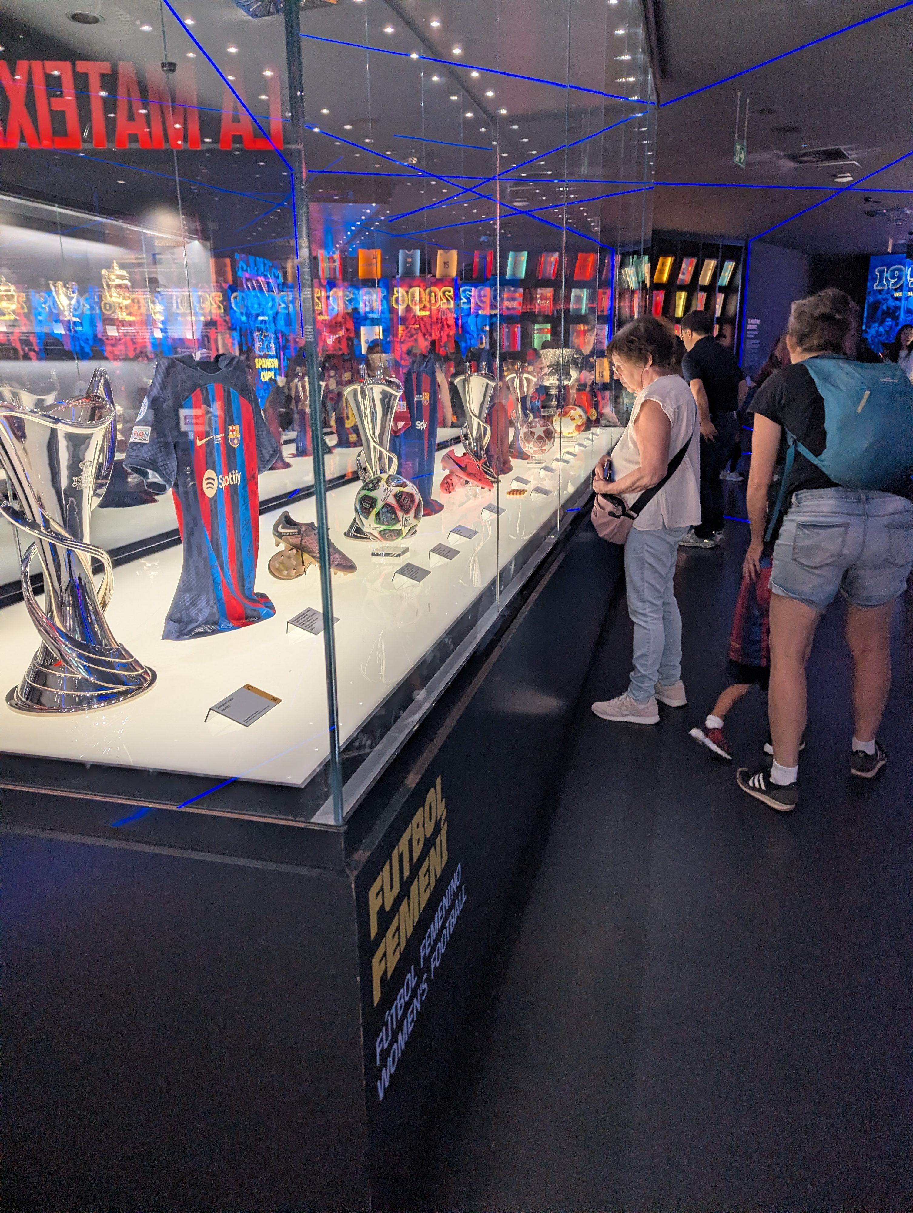 A museum display case of trophies and jerseys labeled Futbol Femení, with visitors looking on