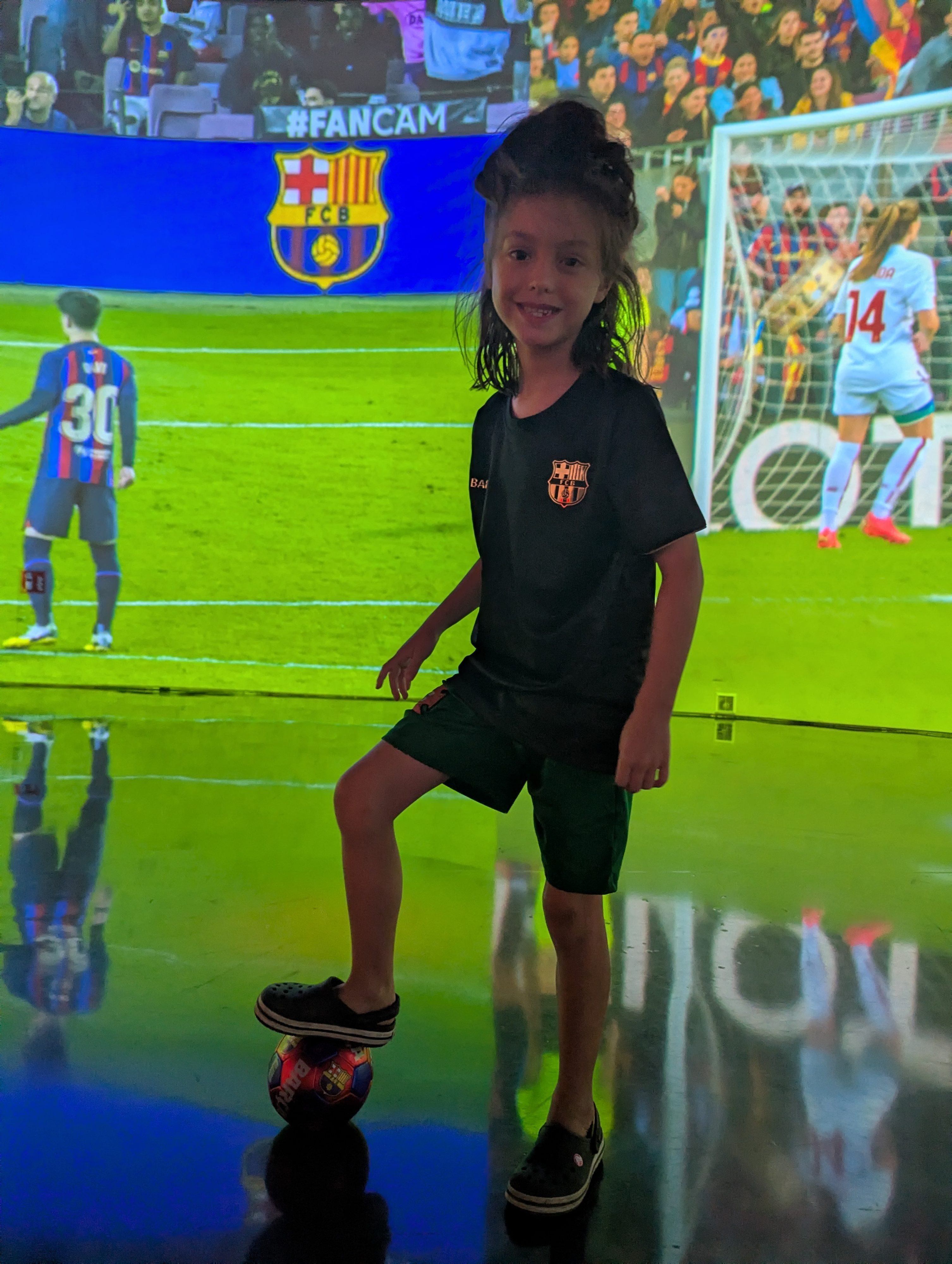 Remi with her foot on a soccer ball on a glowing screen showing a Barça women's match, goal and players behind her