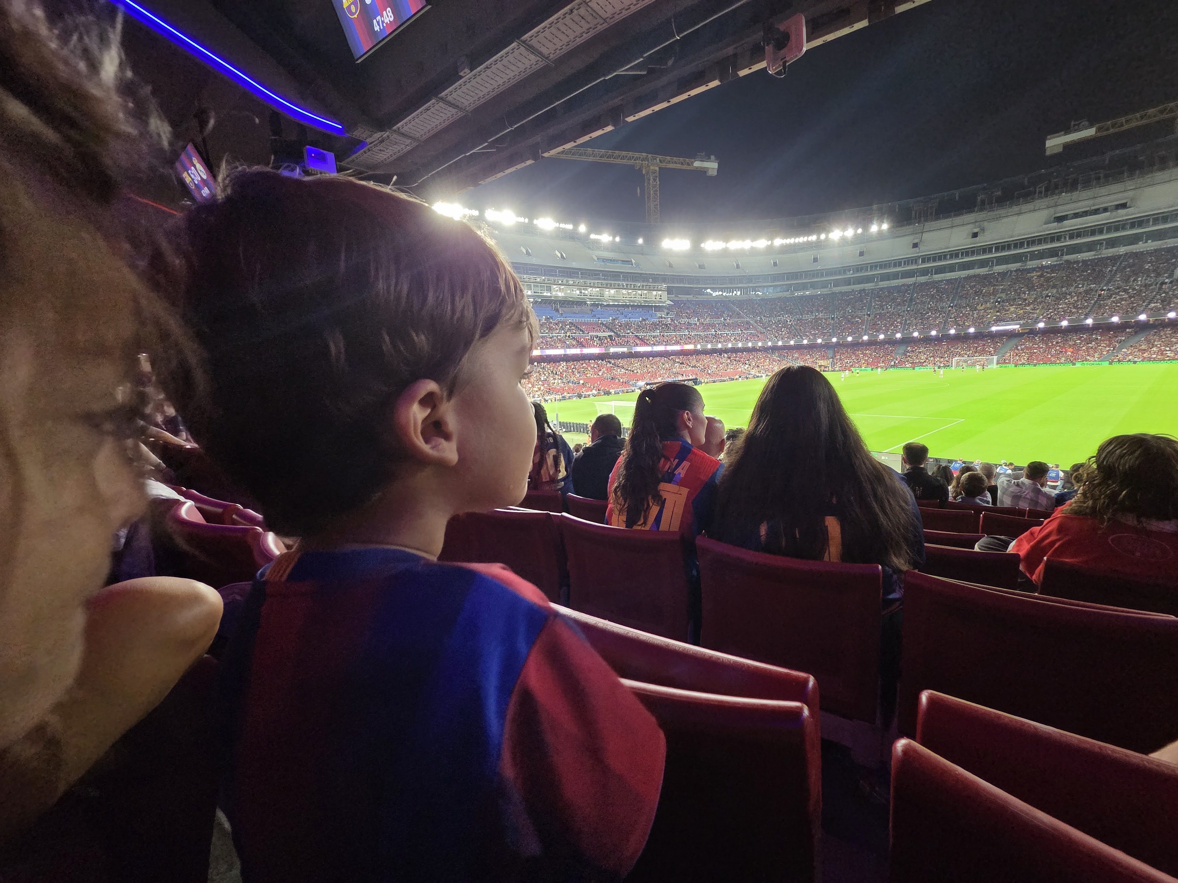 Arlo's head in the foreground in a Barça jersey, looking out over the bright lights and green pitch of the stadium from our seats