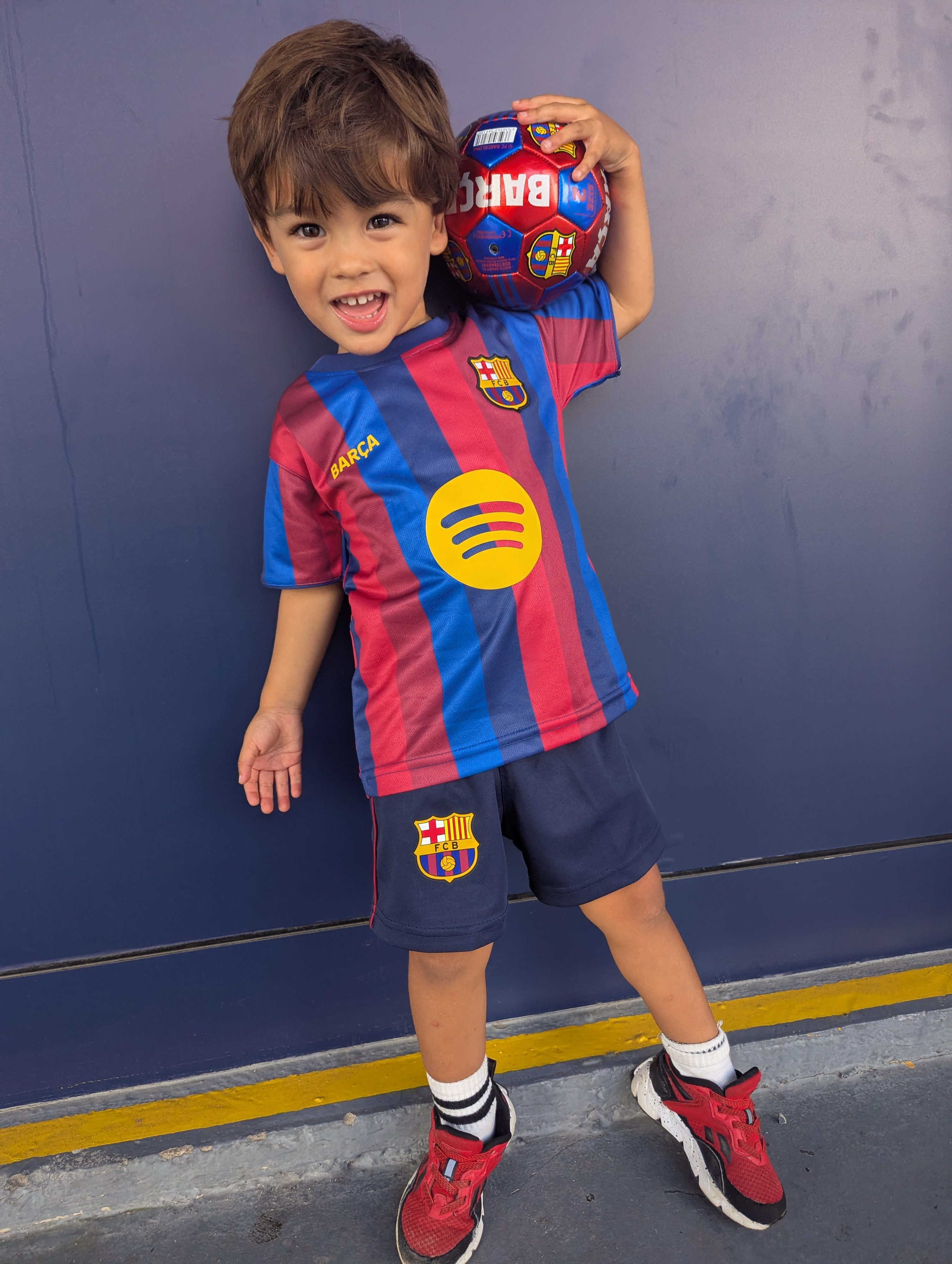 Arlo grinning in a Barça jersey and shorts, holding a Barça soccer ball on his shoulder against a blue wall