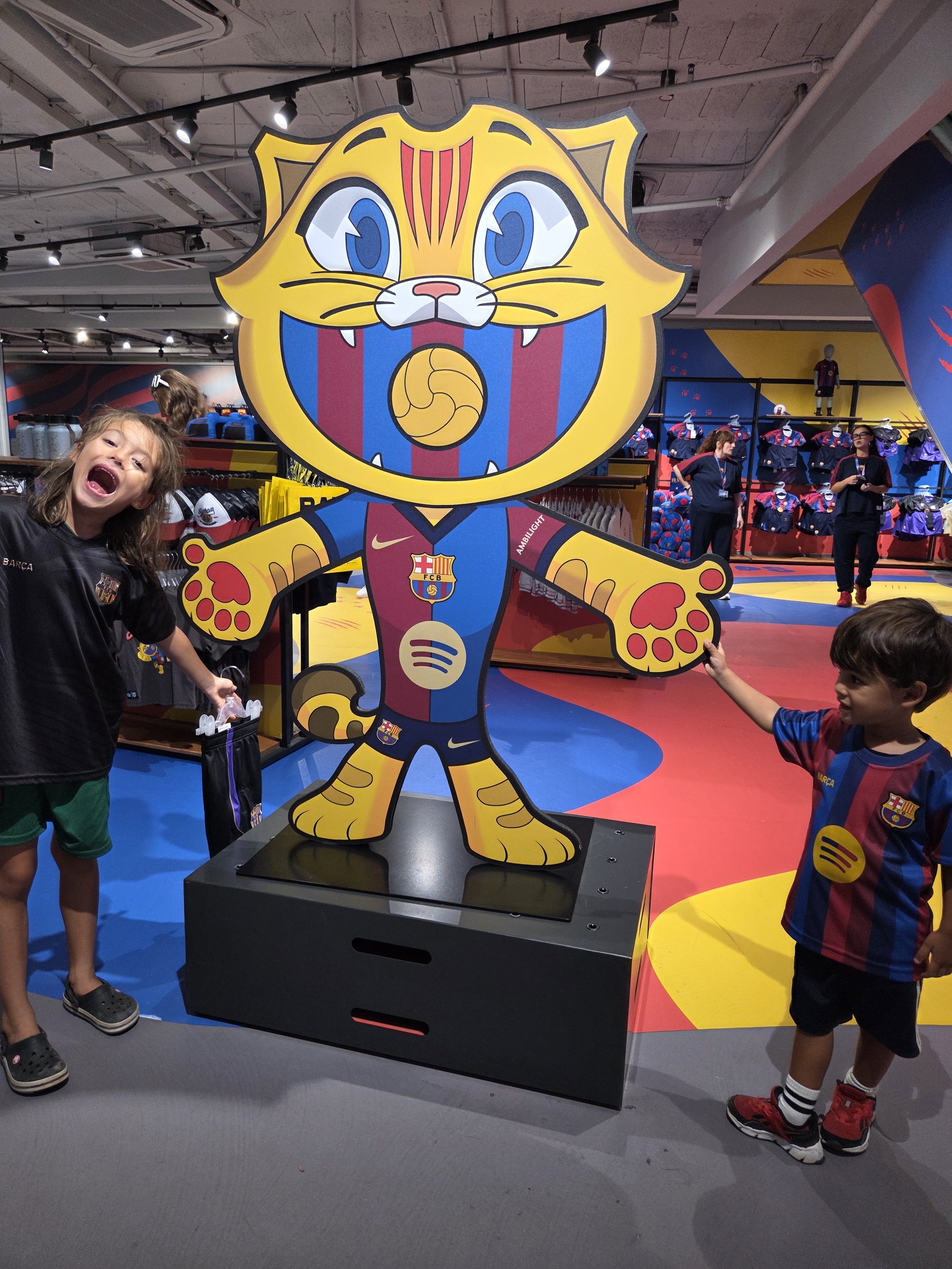 Remi and Arlo posing with a big cartoon cat mascot in a Barça jersey in the stadium shop