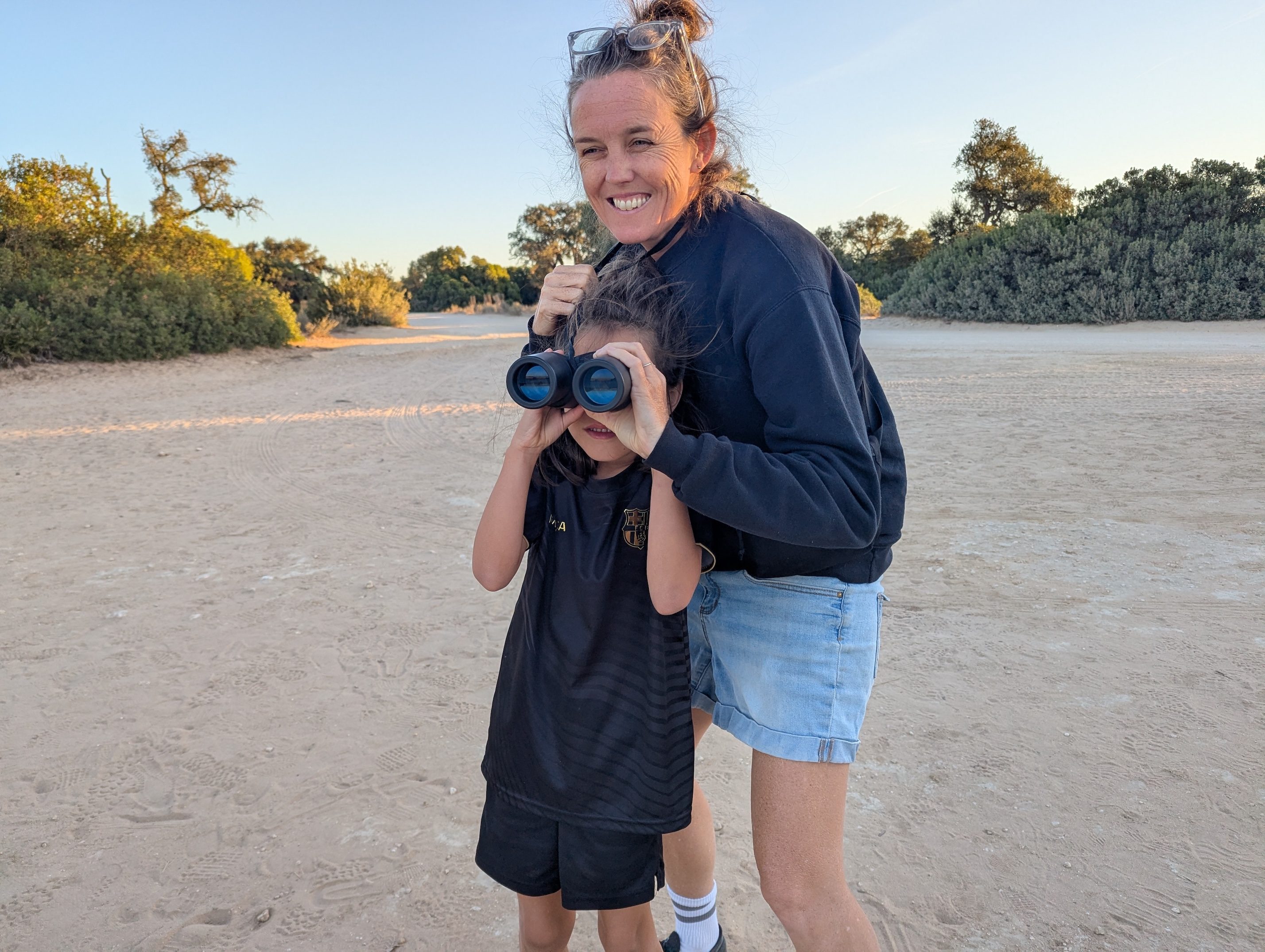 Remi in a black kit holding binoculars to her eyes on a sandy track, with an adult standing behind her smiling, scrubby bushes and morning sky behind them