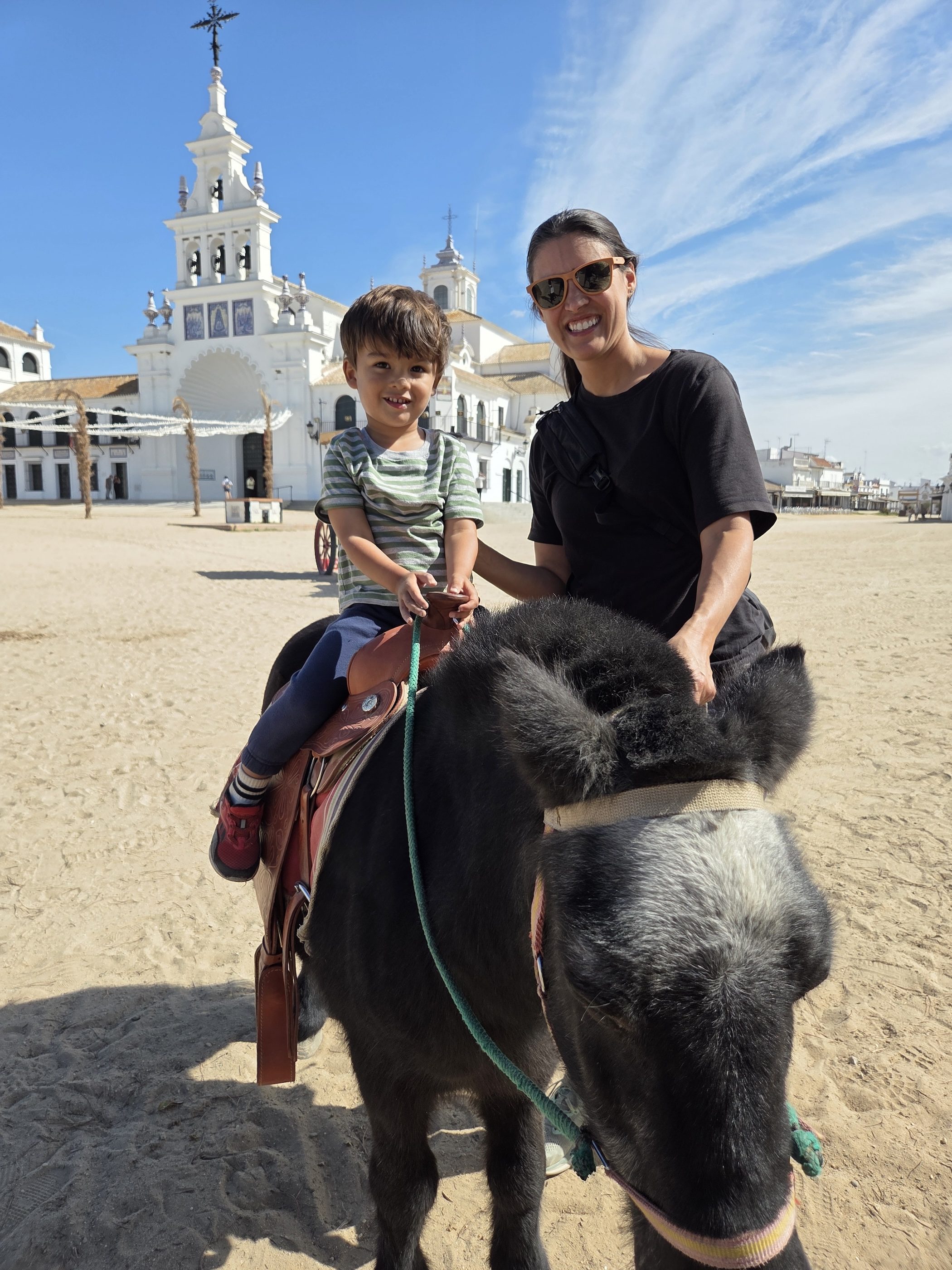 Arlo smiling in the saddle of the same pony, a parent steadying him, the white church behind