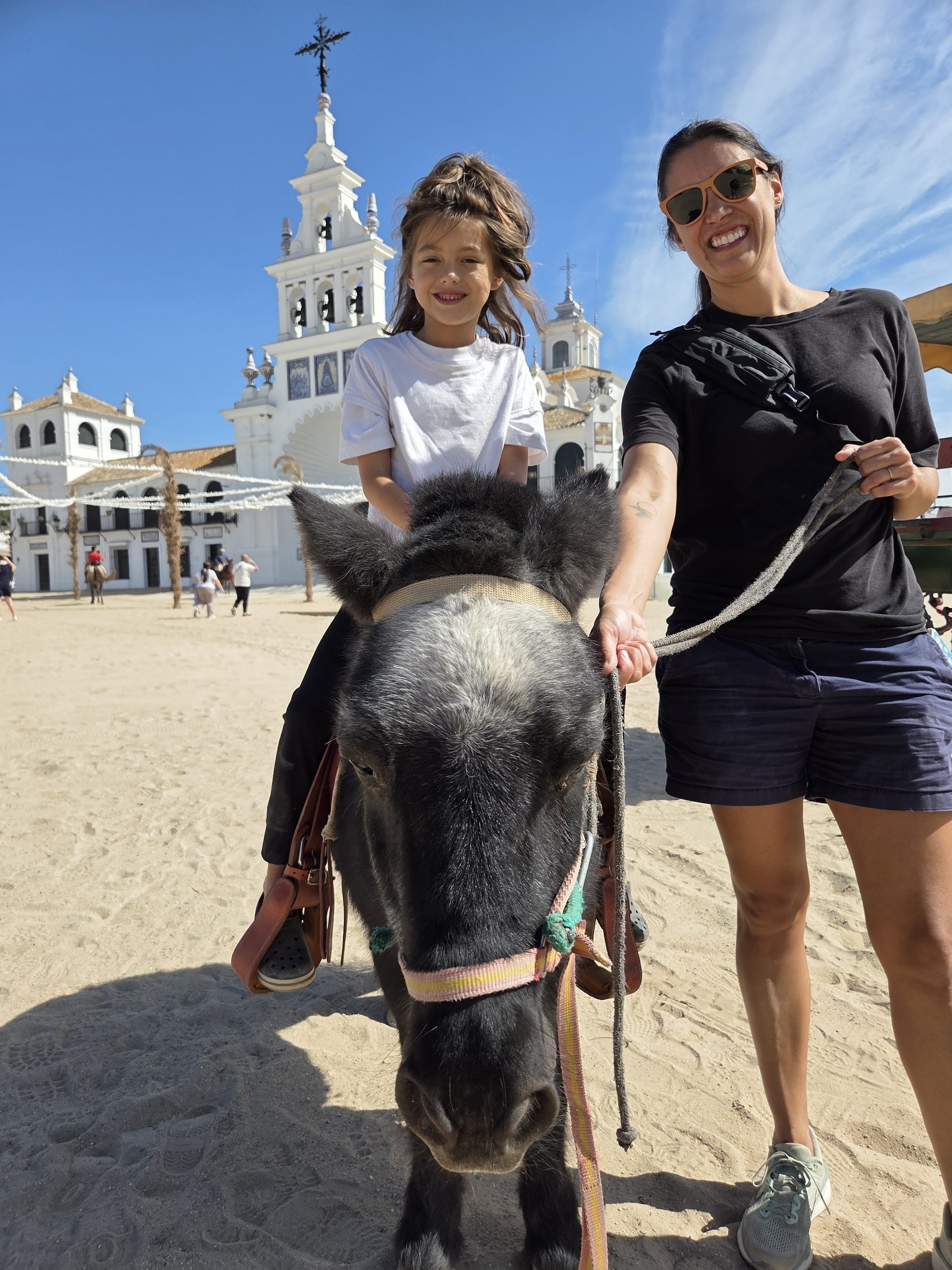 Remi grinning on a small dark pony in the sandy square of El Rocío, led by a parent, with the white church tower behind them
